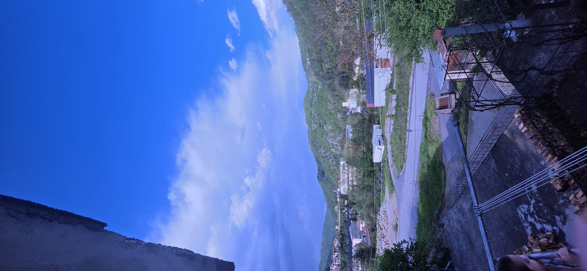 The image presents a view in Rudozem, Bulgaria. A mountain or steep hill, covered in green vegetation, dominates the background. The road winds along the base of the hill, and a few buildings and homes are visible. The sky is mostly blue with some clouds. In the foreground, there are parts of buildings and metallic structures from where the shot was taken.