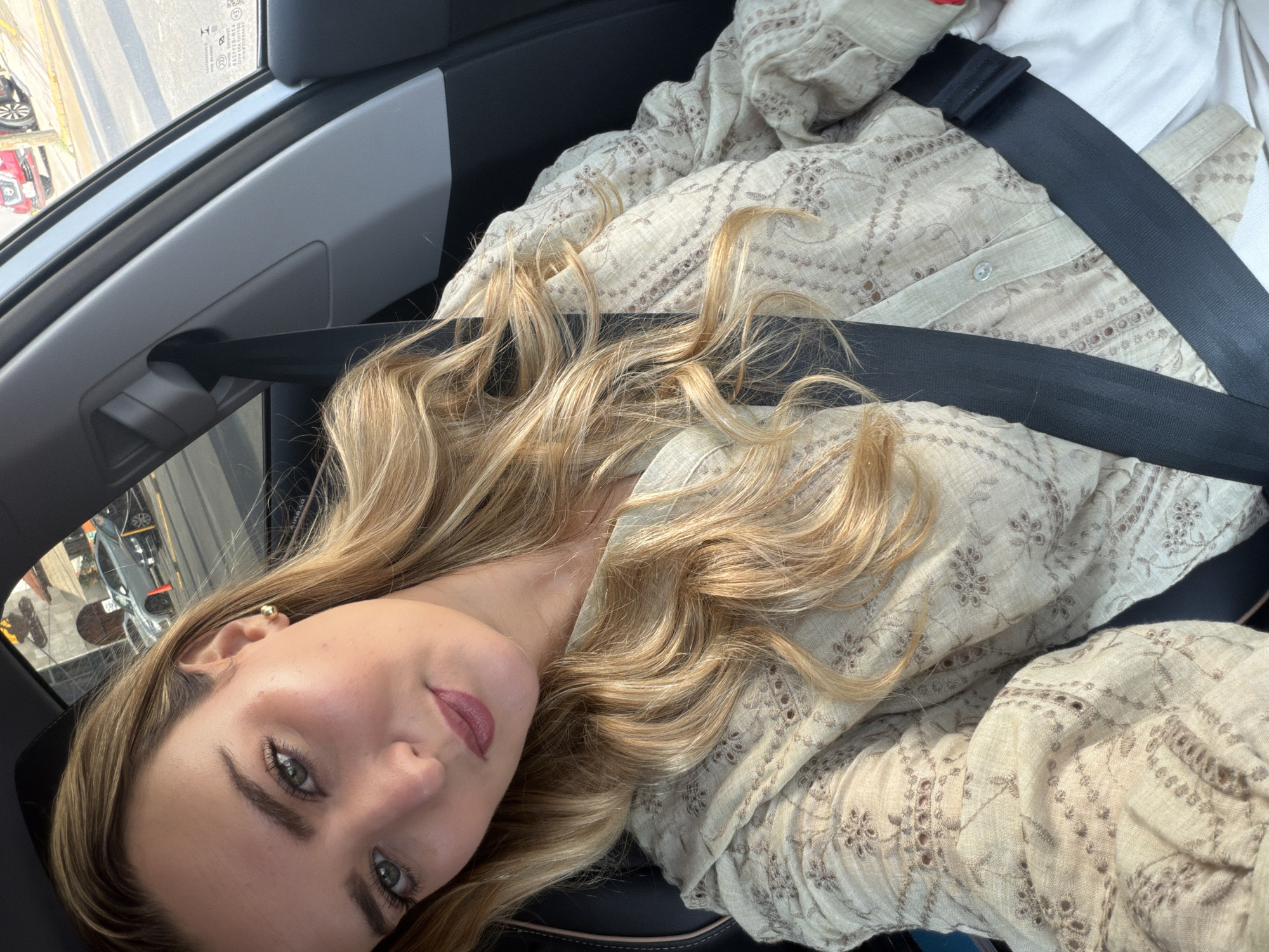 A fair-skinned woman is visible, looking at the camera. She has light blonde, wavy hair and green eyes. She is wearing a beige embroidered shirt and a seatbelt. The scene is inside a car, the interior is black and grey. Through the car window on the left, urban features are visible, including vehicles and buildings, suggesting a street in Puebla, Mexico.