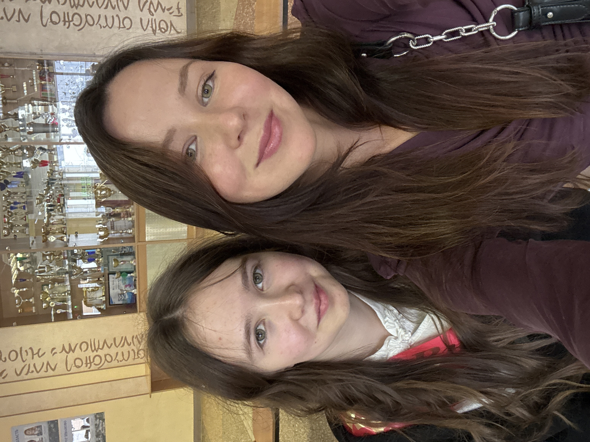 Two women, one older and one younger, are posed closely together for a selfie. Both have long brown hair. The older woman is wearing a plum-colored top, and a black purse chain is visible on her shoulder. Behind them is a trophy case displaying numerous trophies. A wall with text written in Russian is also visible. A sign reads "Ivanovo" and "oblast" which translates to region. This indicates the photograph was taken in the city of Ivanovo, Russia.