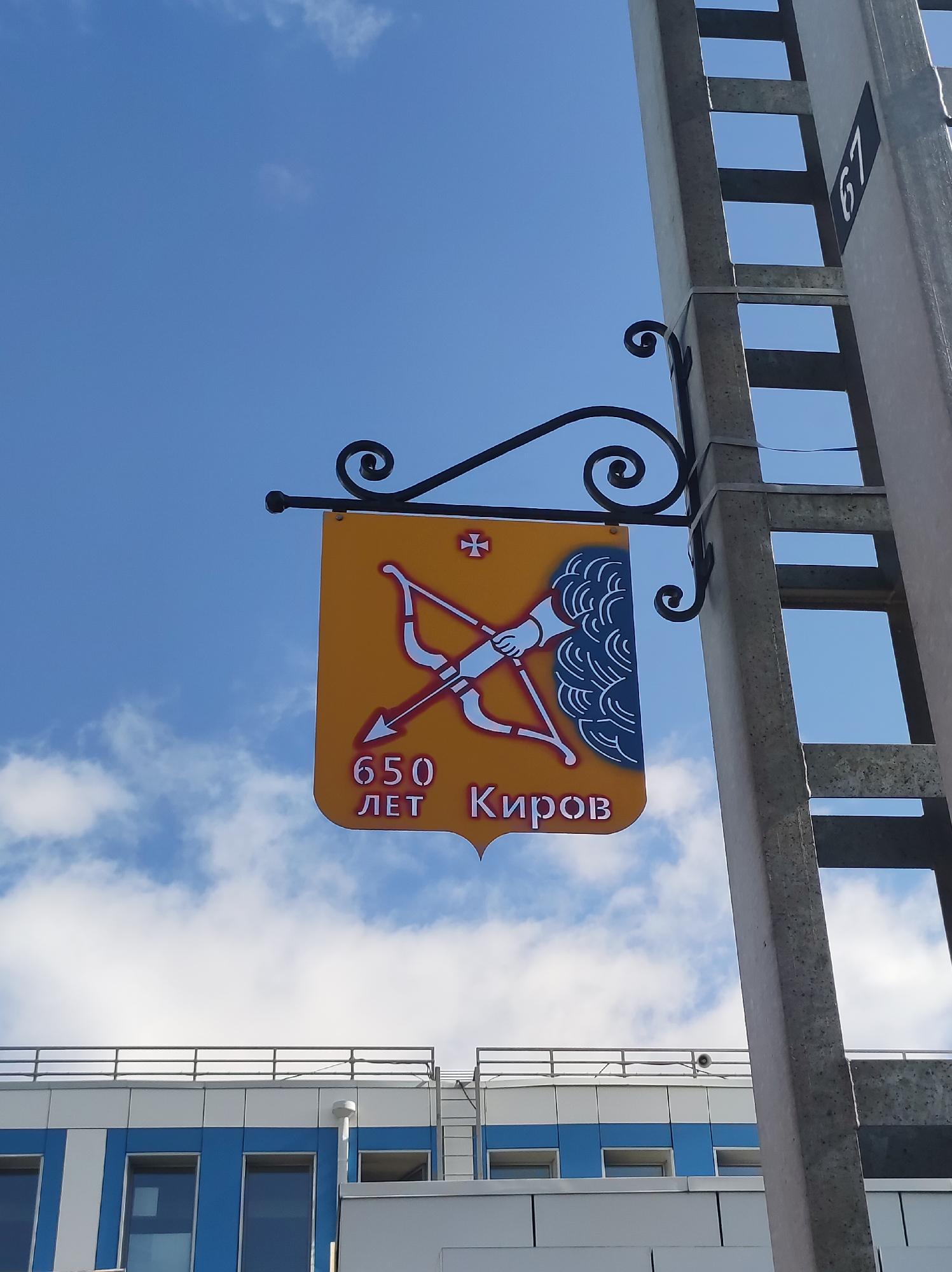 The image shows a yellow sign hanging from a metal pole. The sign has a depiction of a bow and arrow, and the text "650 ЛЕТ КИРОВ" which translates to "650 Years Kirov". The sign is attached to a pole with a black wrought iron bracket. To the right of the sign, a grey metal pole with visible ladder rungs is present. A black rectangular plate with the number "67" is attached to this pole. In the background, a blue and white building with windows is visible under a sky with scattered clouds. This sign likely commemorates an anniversary of the city of Kirov, Russia.