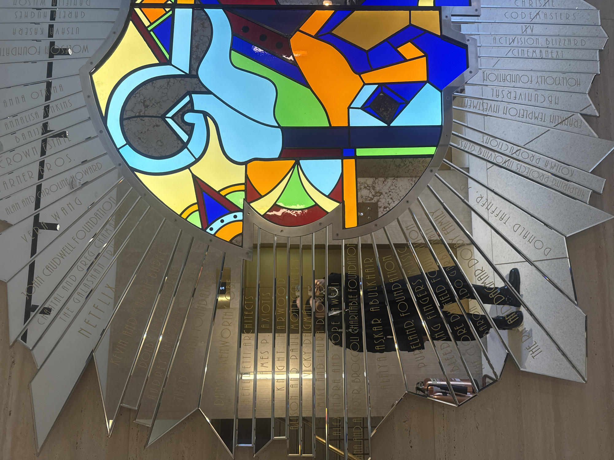 A large, modern stained-glass window, resembling a crest or shield, forms the centerpiece of an elaborate indoor display. The stained glass features abstract, geometric patterns in vibrant hues of blue, yellow, orange, green, and red, with a prominent light blue swirling element. Surrounding the central artwork are numerous silver, highly reflective plaques arranged in a radial, sunburst pattern.
These faceted plaques bear engraved names of individuals and organizations, indicative of a donor or sponsor recognition wall. Visible names include "NETFLIX," "WARNER BROS.," "ACTIVISION, BLIZZARD / CINEMANEXT," "DOLBY," "KOJIMA PRODUCTIONS," "VERA WANG," "CROWN ESTATE," "FRANKLIN TEMPLETON INVESTMENT," and "NBCUNIVERSAL," among many others.
The reflective surfaces of the plaques mirror parts of the immediate surroundings, including a light-colored floor, a person's lower legs and black boots, and a metallic object possibly a fire extinguisher. The reflection of the person suggests they are standing on a staircase or elevated platform. The overall setting, with its modern design and donor wall, is characteristic of a contemporary public building, museum, or corporate lobby, located in London, United Kingdom. The lighting highlights the intricate details and colors of the stained glass, creating a visually striking and dynamic installation.