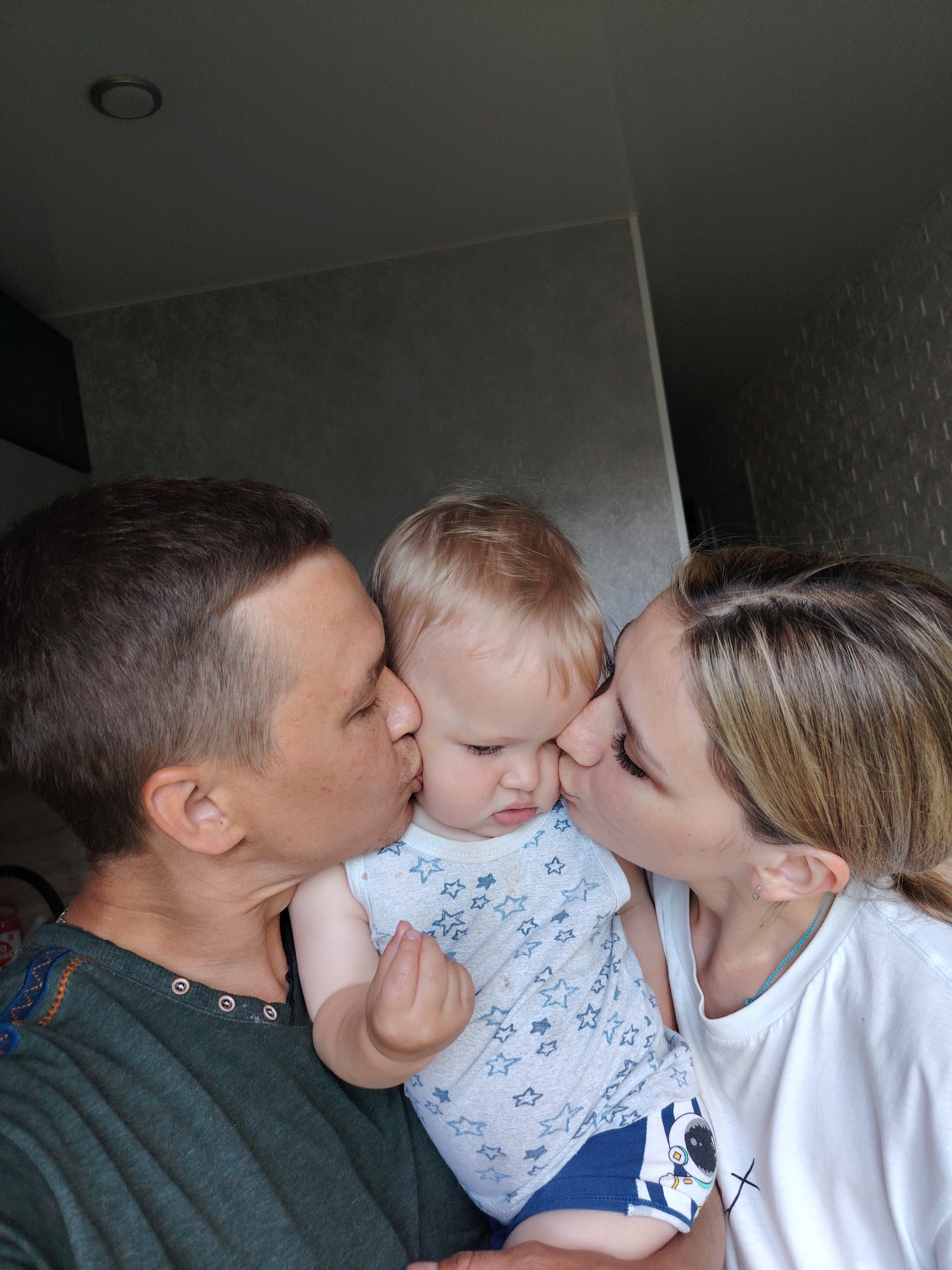 A high-angle indoor photograph captures two adults, a male and a female, simultaneously kissing an infant on opposite cheeks. The scene is set within a residential interior, potentially in Chaikovsky, Russia. The male on the left, with short dark brown hair and a faint stubble, wears a dark green Henley-style shirt featuring copper-colored buttons and embroidered blue and orange geometric patterns on the shoulders. He is shown kissing the infant's left cheek. The female on the right, with blonde hair styled back and prominent eyelashes, wears a white short-sleeved t-shirt with a small black 'X' graphic near the bottom right. She is seen kissing the infant's right cheek. The infant, positioned centrally and held by both adults, has short blonde hair and wears a light grey sleeveless onesie adorned with blue star patterns, over blue shorts with an astronaut graphic on the left leg. The infant displays a slightly frowning expression, looking downwards with pursed lips, appearing either overwhelmed or displeased by the affection. The background consists of dark grey walls with differing textures; the left side shows a darker, possibly furniture or cabinet surface, transitioning to a lighter textured grey wall, while the right side features a textured grey wallpaper with a subtle patterned effect. The dark grey ceiling in the upper left corner contains a single recessed circular light fixture.