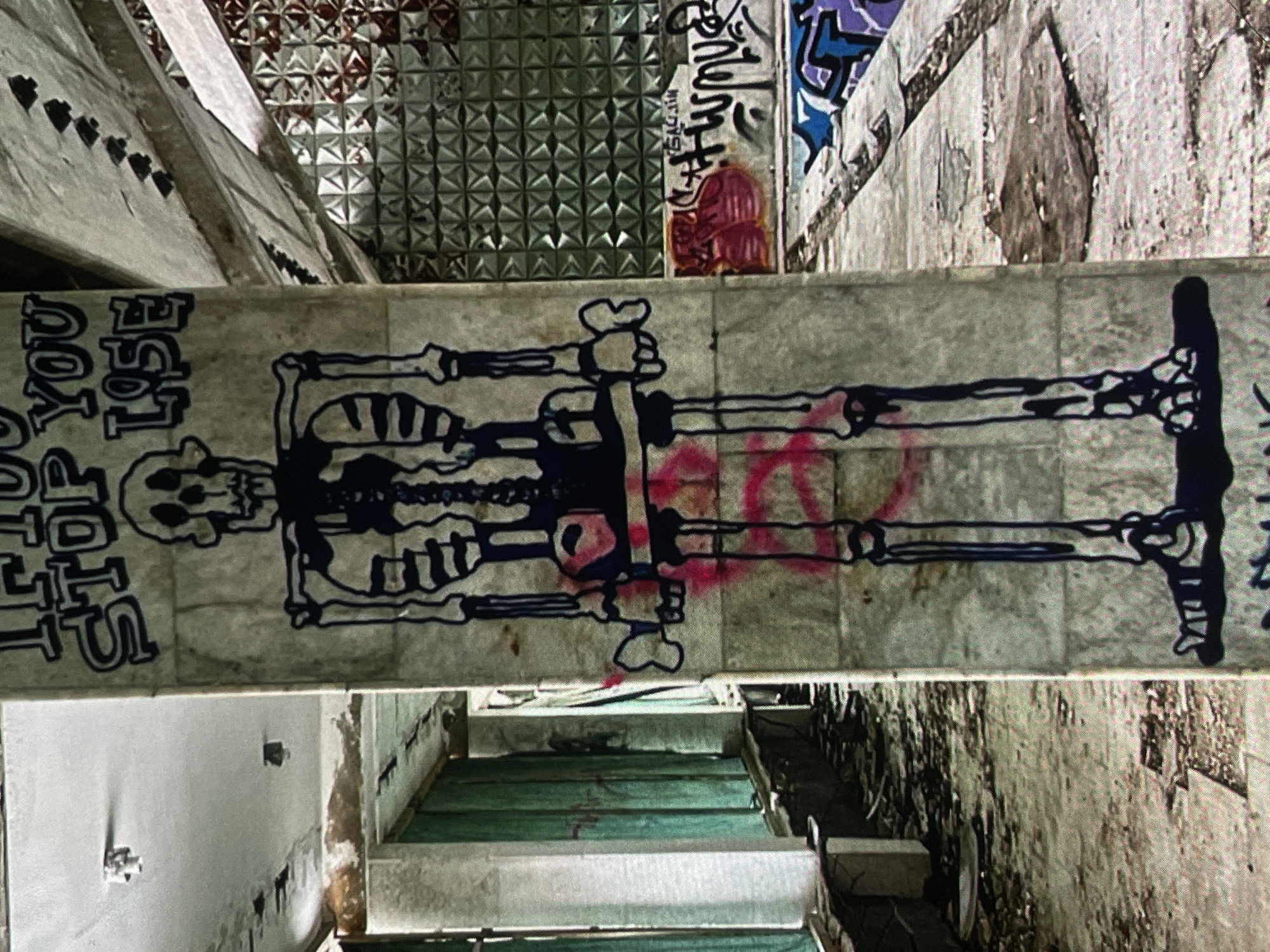 The image captures an interior view of a dilapidated structure, likely an abandoned building, in the city of Harmanli, Bulgaria. Dominating the central focus is a large, crudely drawn skeleton graffiti in black ink with pink accents on a fluted concrete column. Above the skeleton, the text "IF YOU STOP YOU LOSE" is spray-painted in bold, blocky lettering.
The environment is one of decay and neglect. The floor is littered with debris, including dirt, leaves, and fragmented materials. To the left, vertical green tarpaulins or curtains hang in an open doorway or corridor, partially obscuring the view further into the building. The walls and ceiling show signs of water damage and deterioration, with peeling paint and exposed concrete.
To the right of the central column, a wall is covered in more graffiti, including abstract shapes and tags. A distinctive patterned glass or plastic wall with a geometric design is visible in the background, its texture creating a reflective and fragmented appearance. The overall impression is one of an urban decay scene, marked by graffiti art and the passage of time. There are no people visible in the image.