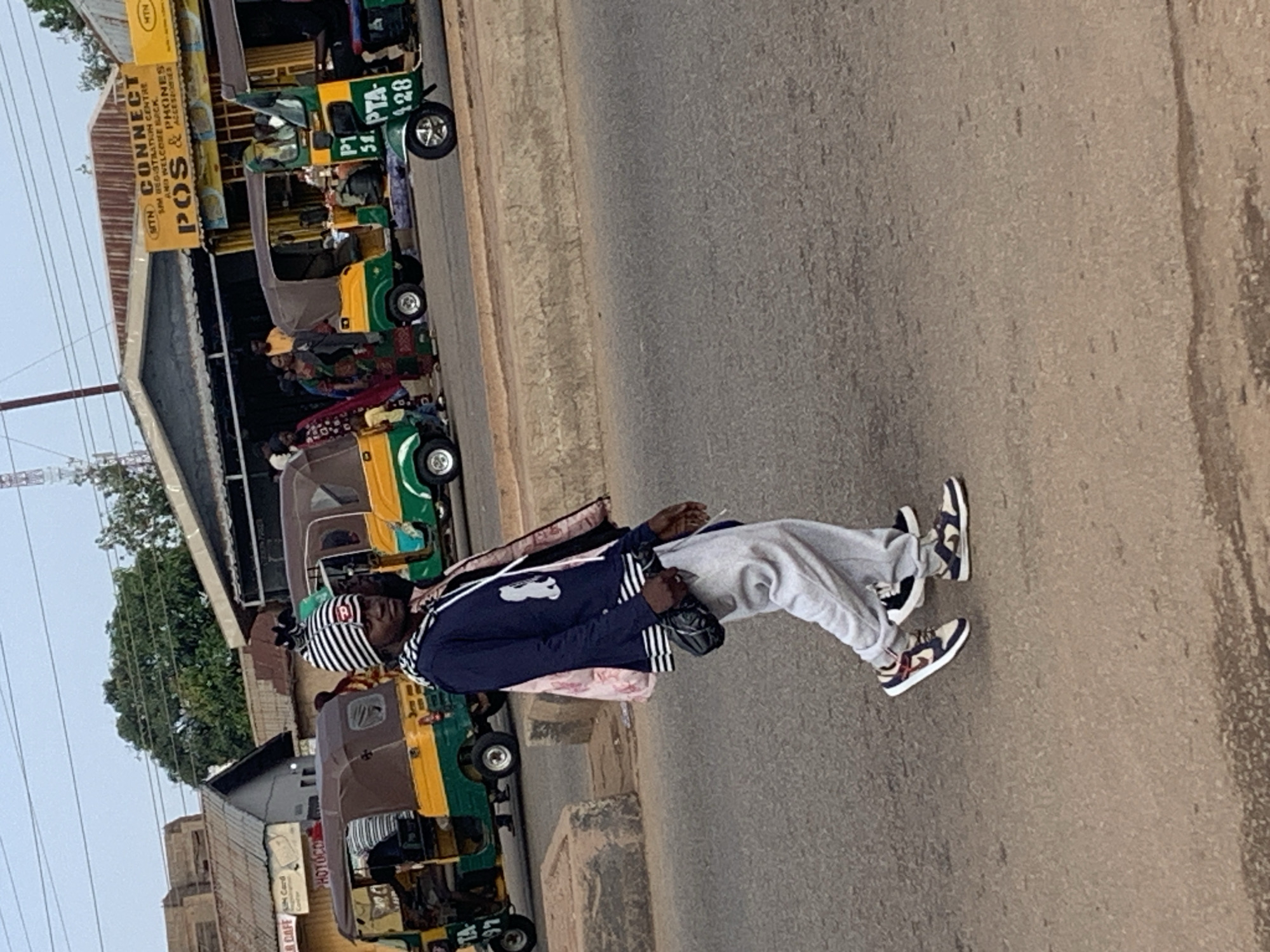 This image depicts a street scene in Gura Top, Nigeria, possibly near a marketplace or transport hub. In the foreground, a person wearing a striped beanie, a dark blue hoodie with a white graphic, light grey sweatpants, and sneakers walks towards the right of the frame. They are carrying a black bag slung across their chest and a light-colored bag over their left shoulder. Behind them, several auto-rickshaws, commonly known as "keke napep" in Nigeria, are parked or in motion. These vehicles are brightly colored, primarily yellow and green. Some of the keke napep have passengers visible inside. In the background, buildings with corrugated metal roofs line the street, one of which displays a yellow sign with text that appears to be a business advertisement, including "MTN CONNECT" and "POS & PHONES ACCESSORIES." The ground is paved with asphalt. The overall impression is a bustling urban environment with a focus on local transportation and commerce.