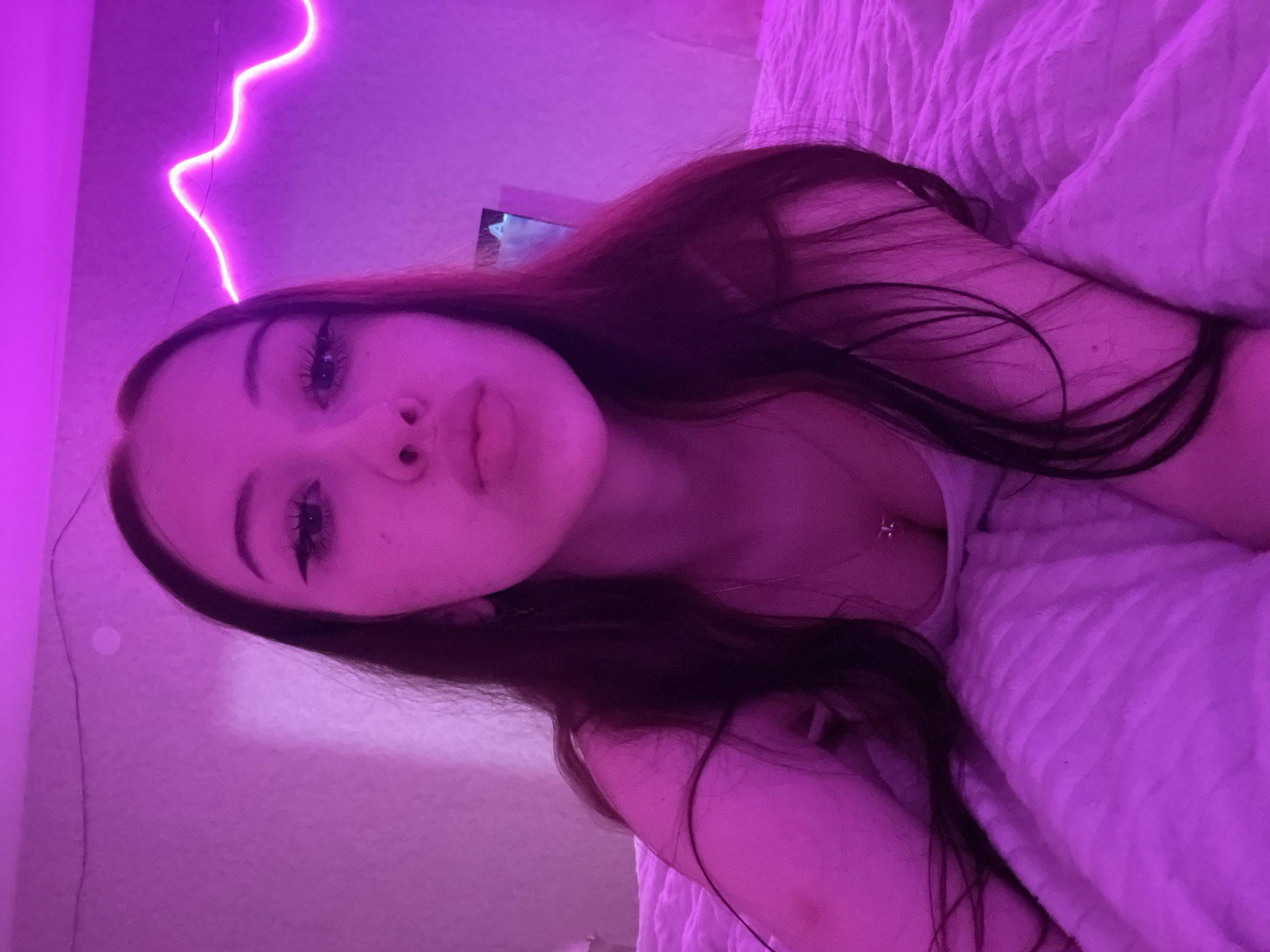 A young woman with long, dark hair and makeup is lying on a bed under pink lighting. She is wearing a light-colored top and a necklace with a pendant. Her lips are slightly pursed, and her eyes are looking towards the camera. A neon light fixture with a wavy design is visible on the wall behind her. The bed is covered with a white, textured blanket. The overall atmosphere is dimly lit and intimate, with the pink lighting casting a warm glow on the scene.