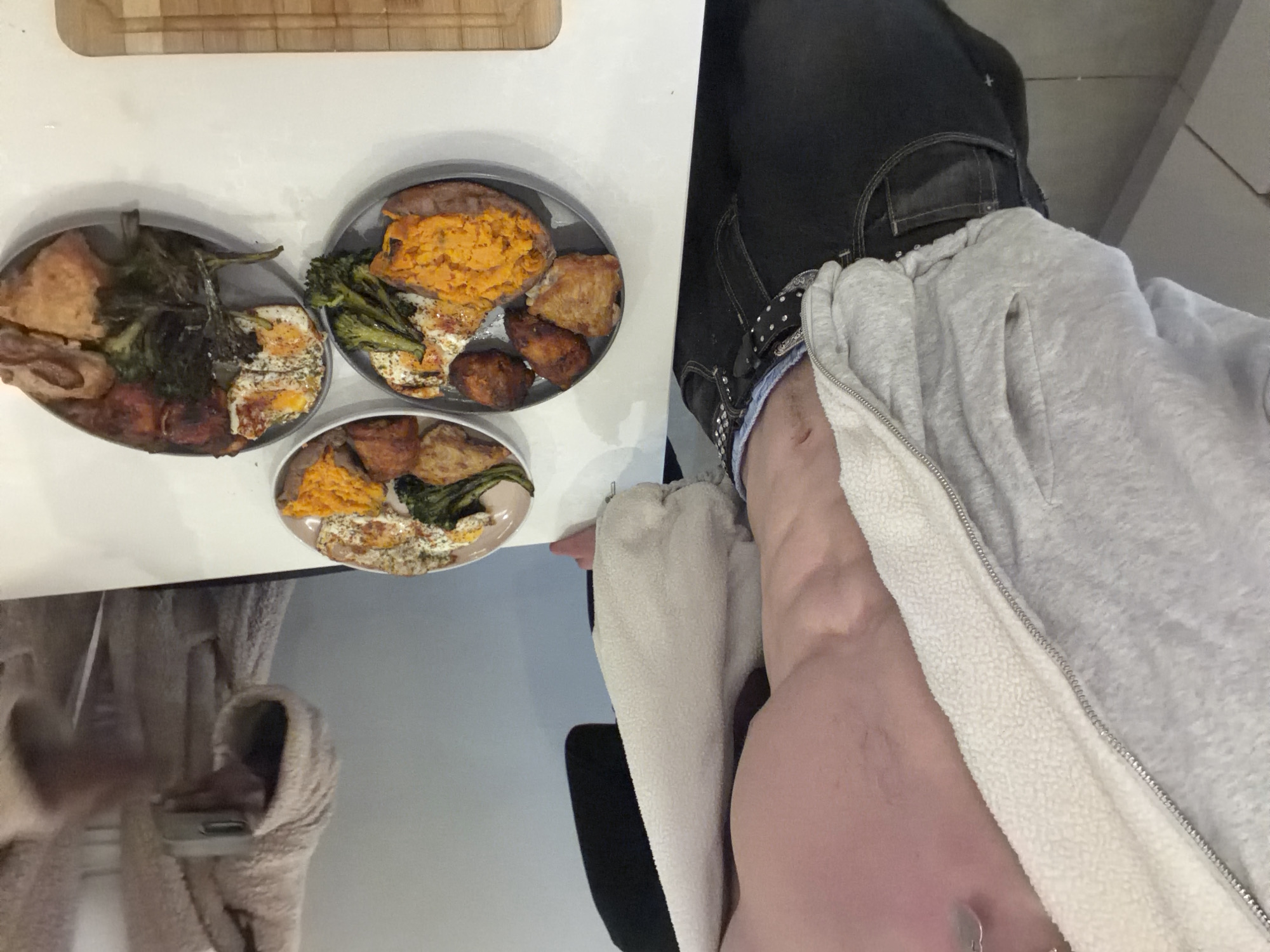 The image, rotated 90 degrees clockwise, captures an indoor scene, likely a kitchen or dining area. Three plates of food are arranged on a white table or countertop. Each plate features a portion of mashed orange vegetable, resembling sweet potato or yam, alongside roasted green vegetables, most likely broccoli or broccolini. Pieces of roasted meat, possibly chicken, are also present on each plate. One plate distinctly shows a fried egg, seasoned with herbs, complementing the meal. A wooden cutting board is partially visible at the top of the frame.
To the right of the food, a person is seated, with a portion of their lower torso and legs visible. They are wearing dark denim jeans with a studded belt and a light-colored (grey and white) fleece-lined zip-up jacket or hoodie. The fabric of the jacket is fluffy on the inside lining. In the blurred background to the left, a large, soft, light-colored object, possibly a cat tree or scratching post, is visible. The overall impression is a home-cooked meal being prepared or enjoyed. No text is visible in the image.