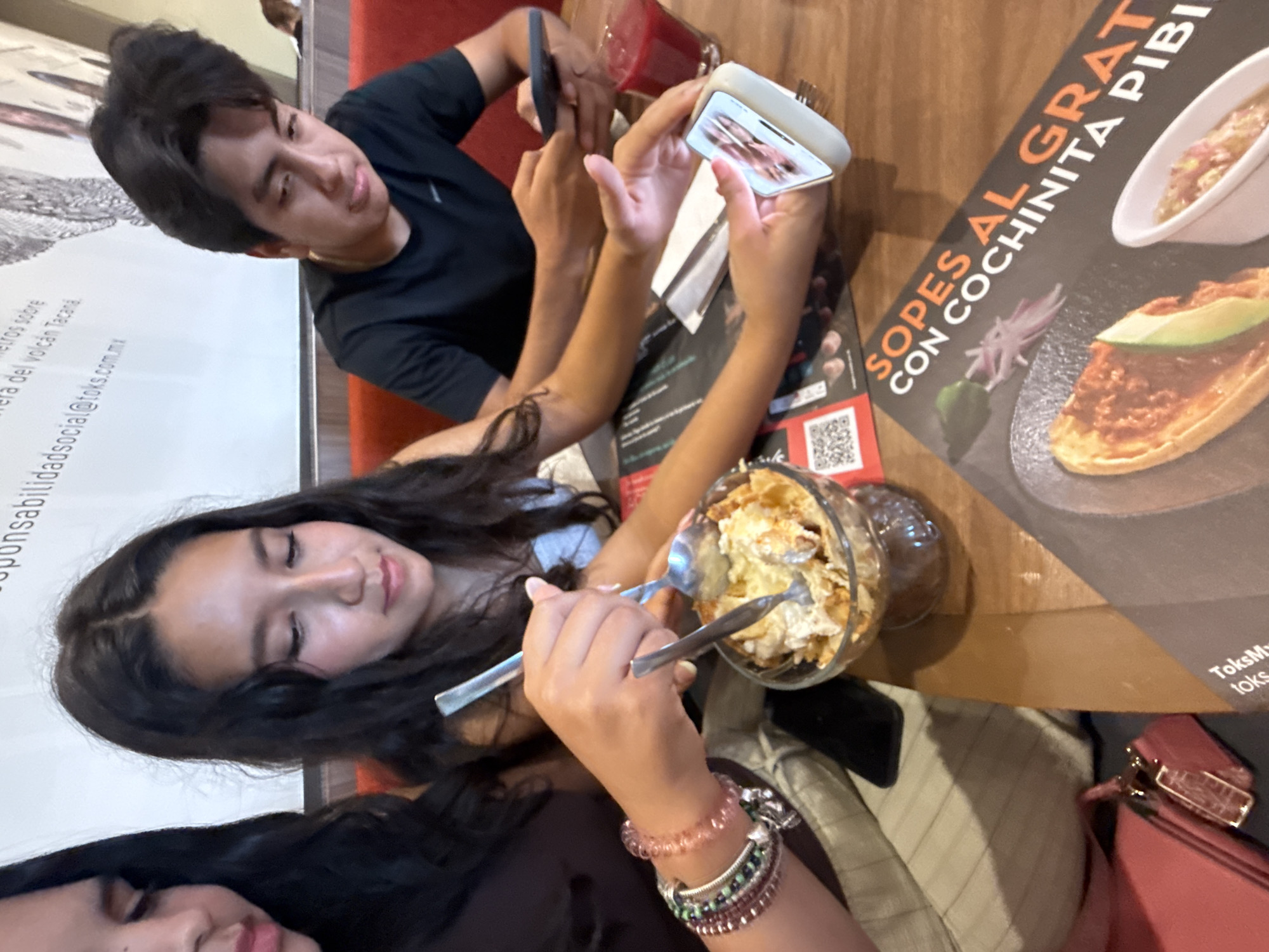 This image depicts a table at what appears to be a restaurant setting in Puerto Marqués, Mexico. Two people are present; a young man, wearing a black t-shirt, is taking a photo with his phone. A young woman, wearing bracelets on her wrist, is eating from a glass bowl with a spoon. At the table, there is a menu featuring the words "SOPES AL GRAT" "CON COCHINITA PIBI", another phone, a red drink in a glass, and a pink purse. The restaurant's name is "Toks".