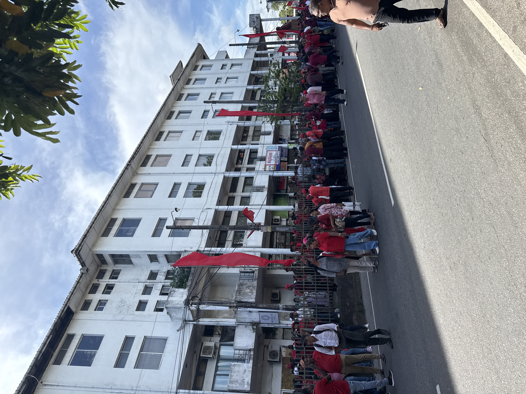 The image captures a street scene in Delkanda, Sri Lanka, showing a large gathering of people, predominantly dressed in red attire, lining a paved road. The perspective is tilted, making the multi-story white building on the left appear at an angle.
In the foreground and extending into the midground, a substantial crowd of individuals is visible, many wearing red shirts, indicating a possible organized event such as a rally or procession. Some red flags are held by participants, while others are affixed to utility poles along the street, reinforcing the impression of a collective gathering. One person in the immediate foreground, on the right side, wears a light brown shirt and dark trousers, holding a mobile phone, and appears to be observing or moving along the street.
The background features a multi-story, white-painted building with numerous rectangular windows. Utility poles with entangled wires run alongside the building. Green foliage from trees is visible in the upper left corner and intermittently along the street. Shadows cast on the road suggest it is daytime, under a clear sky with scattered wispy clouds.
Visible text on storefront signs along the building includes "Highland" followed by what appears to be "Bakery & Sweets". Another sign higher up on the building contains the word "SENANAYAKE" and possibly "Gems". The street itself is asphalt with white painted lines.