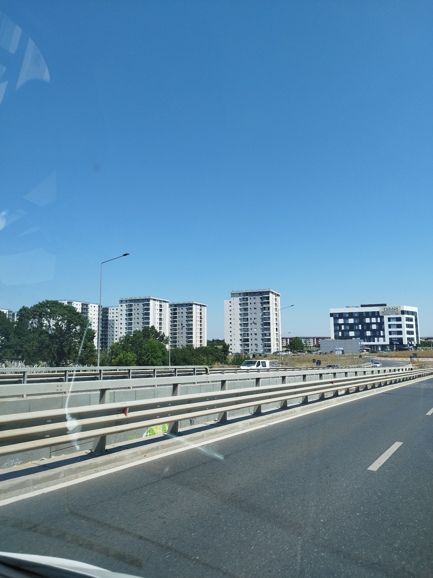 This is a roadside photograph taken on a clear day in Bucharest, Romania. The perspective is from a vehicle moving along an elevated road, indicated by the presence of a concrete barrier with metal railings and guardrails. The road itself is paved asphalt with white lane markings.
In the background, there are several multi-story apartment buildings with a predominantly white and gray facade, featuring numerous balconies. These buildings are situated behind a line of mature green trees. Further to the right, a modern, multi-story office building with a distinct black and white facade stands out. The name "DIMRI" is visible on this building. Another tall, slender apartment building is also present to the left of the office building. Various vehicles, including a white van and several cars, are visible on the road and in the parking areas of the buildings. The sky is a clear, bright blue. The overall scene depicts an urban or suburban area with a mix of residential and commercial architecture.