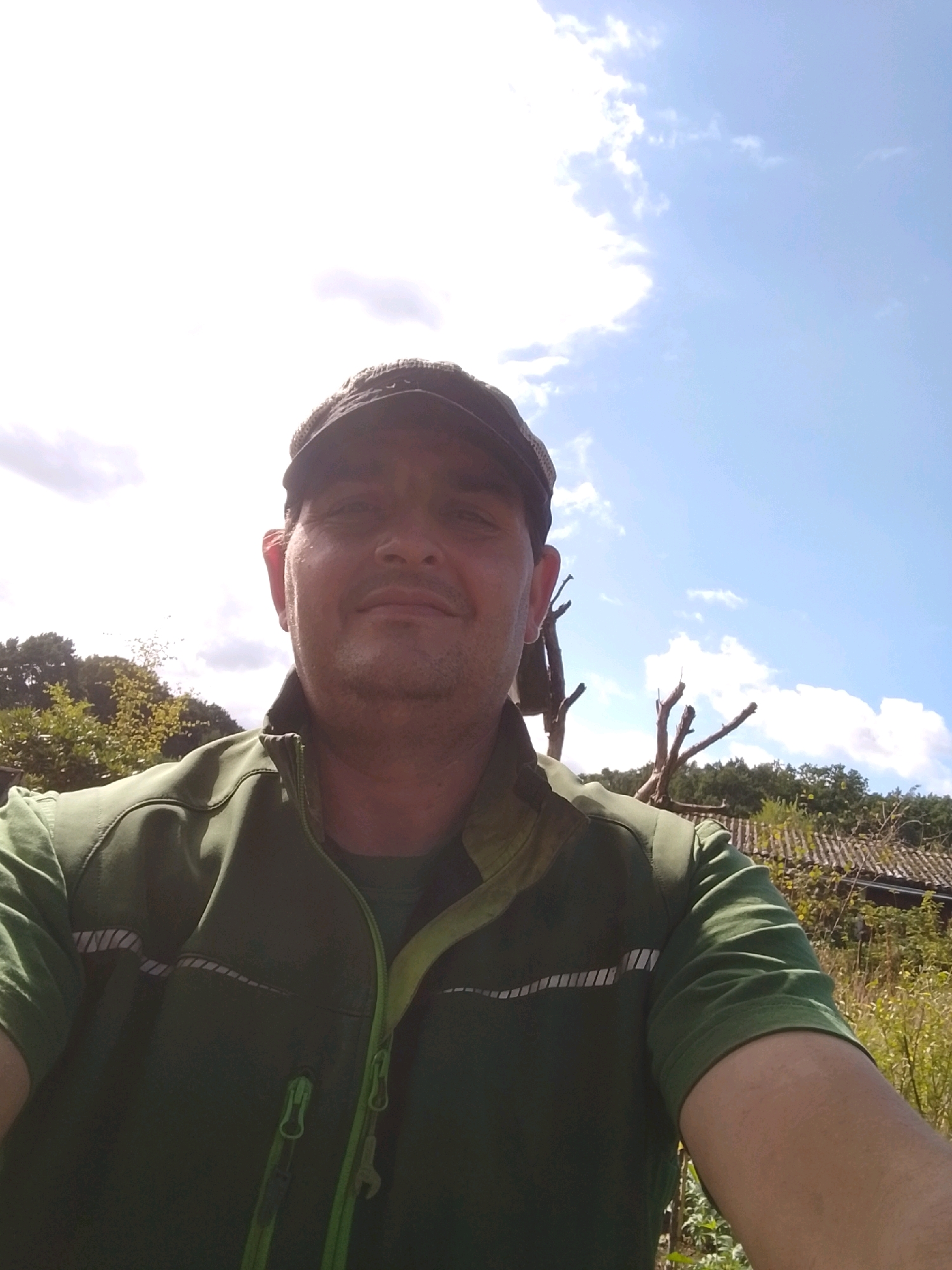 A man in a green t-shirt and a dark green vest with neon green trim is taking a selfie outdoors. He is wearing a dark cap. In the background, there are trees and a cloudy blue sky. The man appears to be in a rural or semi-rural setting. It is daytime, and the sun is bright.