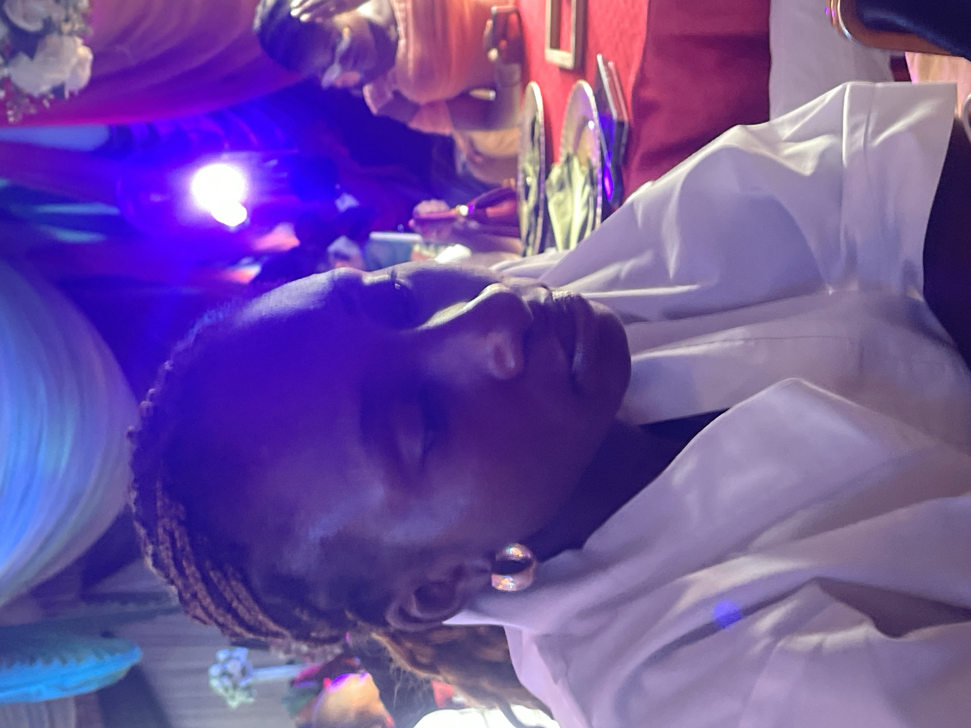 A close-up, tilted, indoor photograph shows a dark-skinned woman with closed eyes and braided hair, wearing a white garment and gold hoop earrings. A bright blue light illuminates the left side of her face. In the background, there is a blurry scene of a table setting with plates, cutlery, and other people, suggesting a gathering or event. The location is identified as Jos, Nigeria, based on external metadata not visible in the image itself.