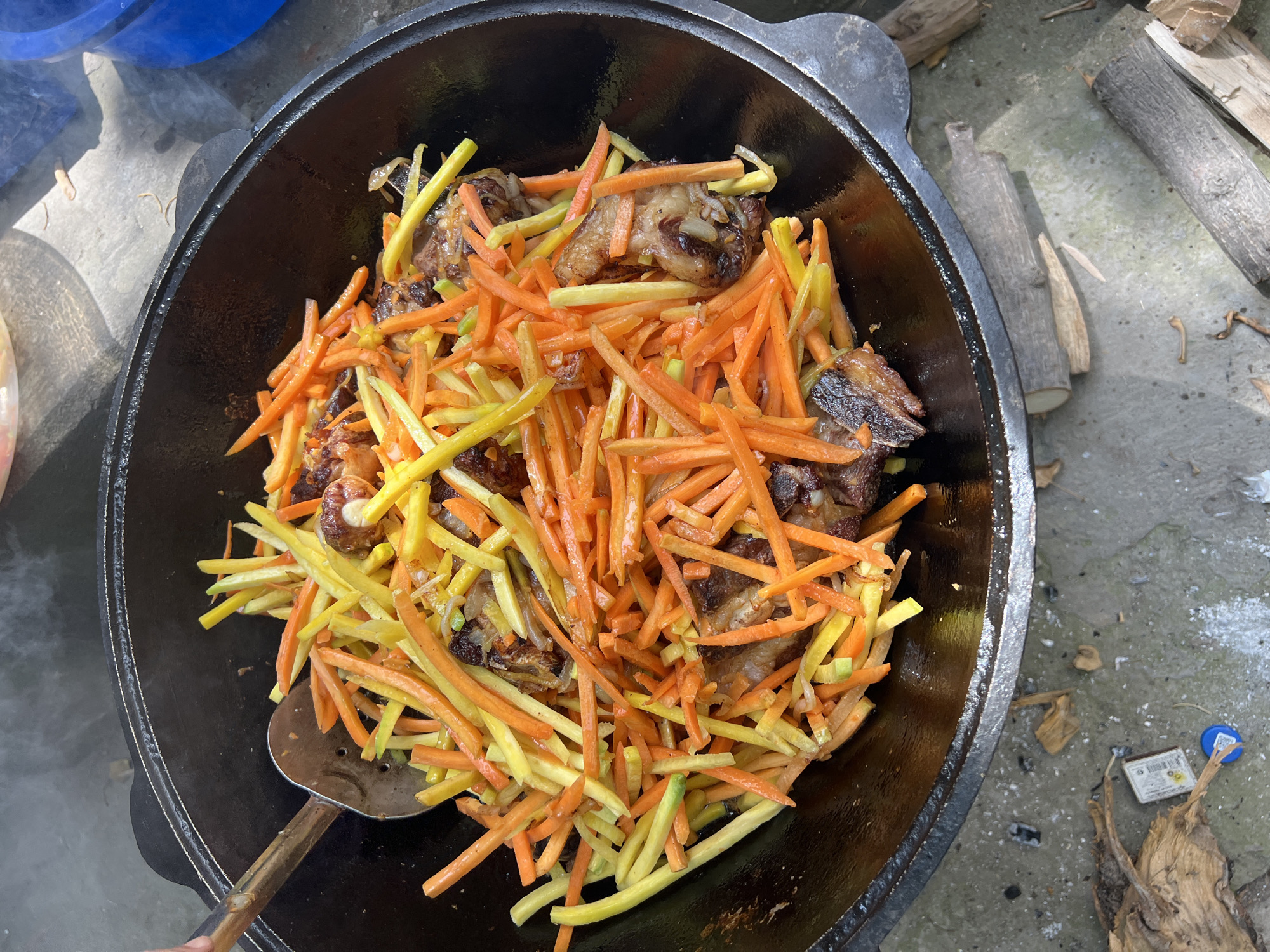 A top-down shot depicts a large, black cast-iron pot filled with a colorful dish of meat and vegetables. The vegetables appear to be julienned carrots in shades of orange and yellow. The meat chunks, browned and interspersed with the vegetables, suggest a stew or stir-fry. A large metal spoon with a wooden handle is partially submerged in the food, indicating it's being stirred or served.
The pot is placed on an uneven concrete surface. To the upper right of the pot, there are several pieces of chopped wood, possibly for cooking. A blue plastic bucket is visible to the upper left. Litter such as a matchbox and small debris are on the concrete surface near the bottom right of the pot. The overall composition gives the impression of outdoor cooking.
Given the food style in the image, it could likely be a food stall within a Bazaar or a roadside setup.