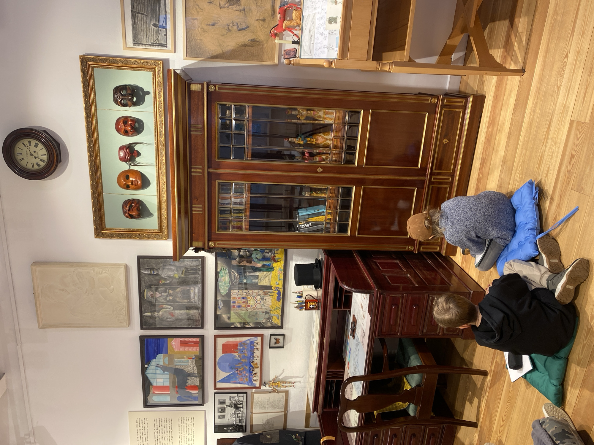 Two children are seated on cushions on a wooden floor in a room filled with art and antique furniture. The setting appears to be a museum, gallery, or a richly decorated private study. The walls are white, and the floor is made of light-colored wood planks.
The child closer to the viewer, wearing a black hoodie, is on a green cushion, seemingly looking at papers or a book. The other child, in a grey sweater and a brown cap, is seated on a blue cushion, facing away from the camera, also focused on an activity on the floor near a large wooden cabinet.
The room is adorned with numerous framed artworks and objects. On the left wall, a round wooden wall clock with Roman numerals shows approximately 11:10 or 11:50. Below it is a tall, framed display containing five intricately carved masks. Adjacent to these, a large, ornate dark wooden cabinet with glass doors showcases items like dolls or figurines and books; one visible book spine reads "БАРХИН" in Cyrillic.
To the left of the cabinet, the wall is covered with various framed pictures, including a light-colored relief sculpture, several paintings with military figures, abstract compositions, and colorful architectural scenes. Below these artworks, a framed document containing Cyrillic text is visible.
To the right of the cabinet is a dark wooden desk with a chair, upon which a black top hat is placed, along with a pencil holder. The desk surface is also covered with papers or designs. Further back, a lighter wooden shelving unit is visible. The overall impression is one of a space dedicated to culture, history, and learning.
