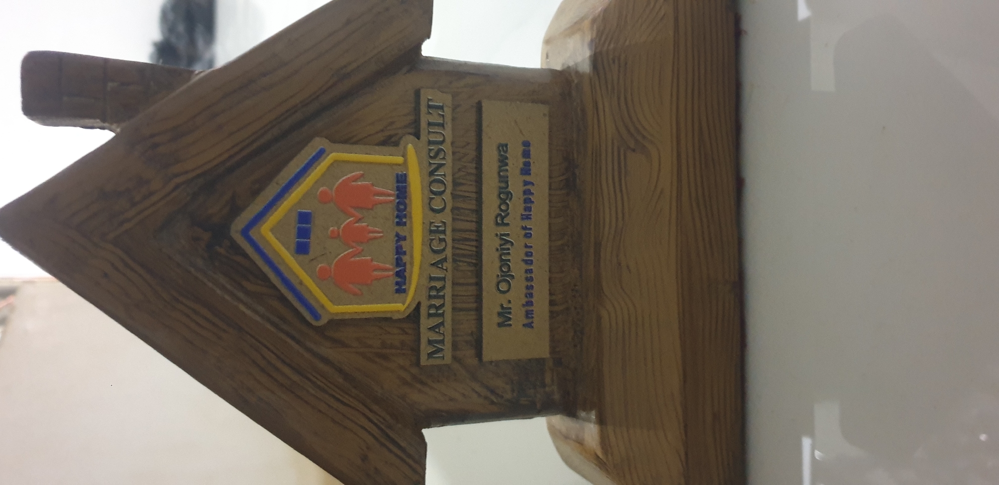 The scene is located in Barden Yana, Nigeria. A brown wooden plaque, carved in the stylized shape of a house with a peaked roof and a small chimney on the right side, stands upright. The wood exhibits a prominent grain texture, suggesting a natural finish. On the front facade of the house, a multi-colored emblem is embedded. This emblem features a yellow house outline with a blue roof segment and three blue square windows. Inside the yellow house outline, three orange, stylized human figures are depicted: two larger figures (adults) flank a smaller central figure (child), with all holding hands. Below these figures, the words "HAPPY HOME" are embossed in blue capital letters. Below the emblem, a rectangular plate contains the dark, embossed capital letters "MARRIAGE CONSULT." Directly beneath this, another rectangular plate displays two lines of dark, embossed text: "Mr. Ojoniyi Rogunwa" followed by "Ambassador of Happy Home." The house-shaped plaque is mounted on a flat, rectangular wooden base, consistent with the house's material and finish. The object rests on a smooth, reflective white surface, likely a table, which shows a slight reflection of the plaque's base. The background is out of focus, appearing as a light, off-white wall or surface, with a darker, indistinct area visible towards the upper right. The lighting is bright and direct, illuminating the front of the plaque.