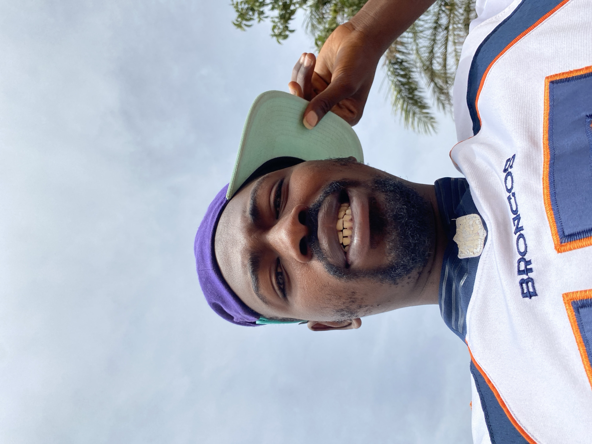 The image shows a man wearing a purple head covering and a Denver Broncos jersey. He is smiling and his teeth are visible. A hand is adjusting the visor of his head covering. The background is a cloudy sky and the top of a palm tree is visible. The image does not provide enough information to determine if it was taken in Barakin Akawo, Nigeria.