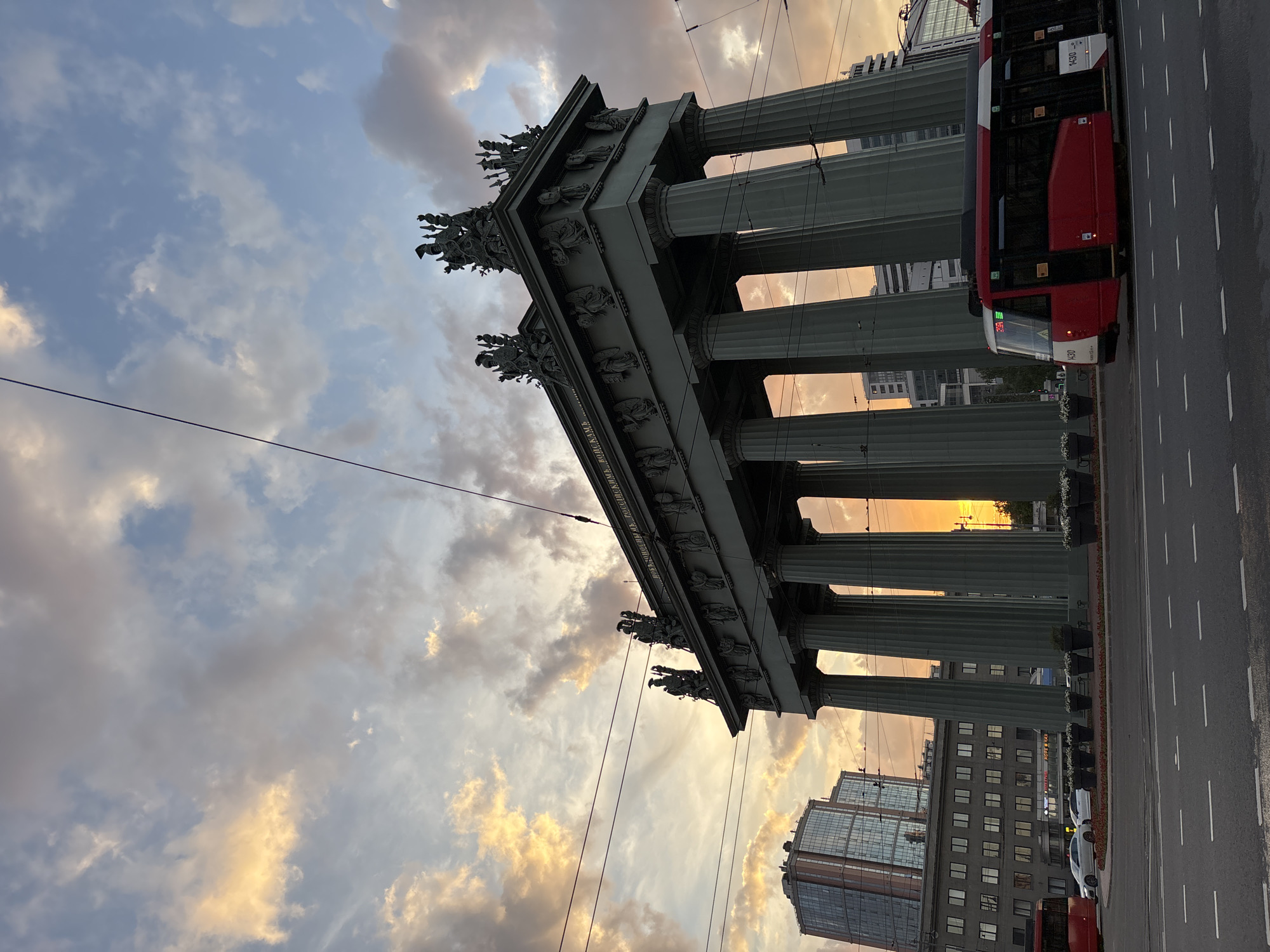 A striking urban scene in Saint Petersburg, Russia, is captured during either sunrise or sunset, bathing the surroundings in a warm, golden light. The focal point is a monumental neoclassical building, likely the Kazan Cathedral, characterized by a grand colonnade of numerous tall, grey columns. Its facade and roofline are adorned with intricate sculptural details and statues of figures. A visible Cyrillic inscription along the frieze reads: "БЛАГОДАРНОЕ ПОТОМСТВО ВЕЛИКОМУ ПОБЕДИТЕЛЮ" (Grateful Posterity to the Great Victor).
The sky above is painted with a mix of blue, grey, and warm golden-orange clouds, illuminated by the low sun on the horizon, which casts a radiant glow through the columns. To the right, a paved city street features a modern red and white tram or trolleybus, marked with the number "1430", appearing stationary. Overhead electrical wires crisscross the sky, indicating urban transport infrastructure. In the background, modern multi-story buildings with glass exteriors provide a contemporary contrast to the classical edifice. Planters with white flowers line the base of the columns along the street.