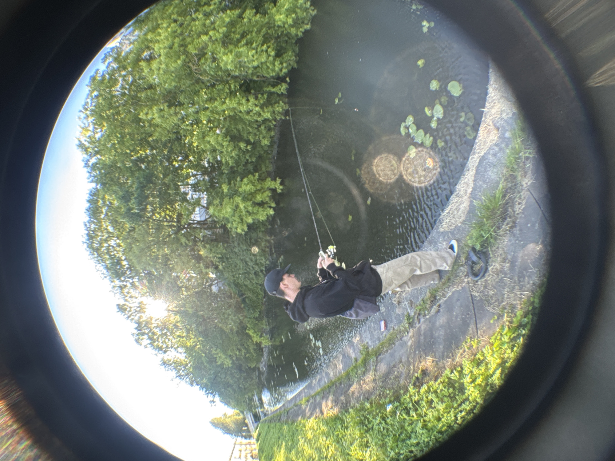 A young male wearing a black baseball cap, black hoodie, and light-colored pants is fishing on the bank of a narrow canal or river. He is holding a fishing rod in his hands, and the line extends into the water. The water has lily pads on its surface, and lush green trees line the opposite bank. The sun is shining brightly, casting reflections on the water and creating lens flares. The image is distorted by a fisheye lens, giving it a wide-angle, curved appearance, and a dark vignette surrounds the scene. The location is identified as being in Saint Petersburg, Russia.