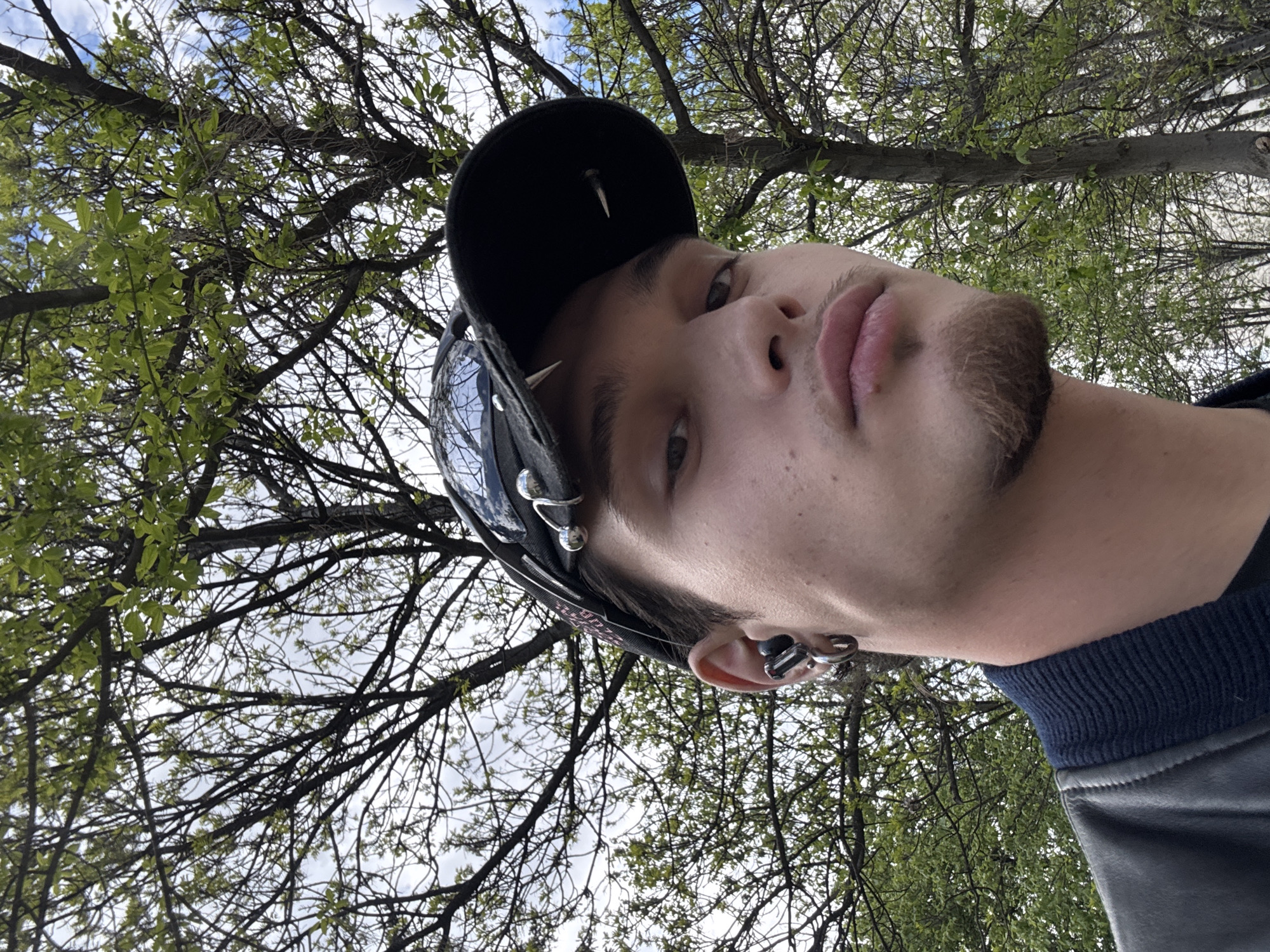 The image features a close-up, upward-angled portrait of a young person, likely male, positioned outdoors under a tree canopy. The individual has light skin, dark eyes, and a short, light-colored goatee and chin beard. They have full lips and gaze directly at the camera with a neutral expression.
They are wearing a black cap adorned with several prominent silver-toned spikes or studs on its brim and potentially on the crown. Multiple silver-toned earrings are visible on their left ear, including several ear cuffs along the helix, a hoop, and a stud in the earlobe. The collar of a dark garment, featuring a ribbed, dark blue or black fabric, is visible below their chin.
The background is dominated by the intricate network of tree branches, many of which are bare, while others are beginning to sprout fresh, bright green leaves, indicating spring or early summer. Patches of light blue sky with scattered white clouds are visible through the foliage, suggesting it is daytime with partially cloudy weather. The perspective is from a low angle, looking up towards the subject and the sky. No discernible text is visible in the image. The location context is Sofia, Bulgaria.