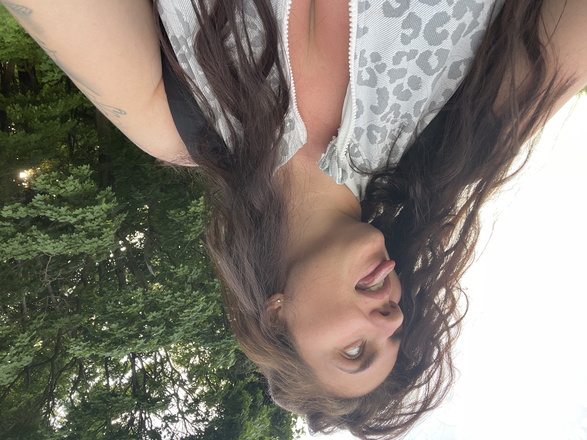 A person is looking up at the camera with their tongue slightly sticking out. They have long, brown hair and fair skin. They are wearing a light gray top with a patterned design. The background consists of green trees and a bright, overcast sky. The location in the city of Ostrov, Russia cannot be determined from the image.
