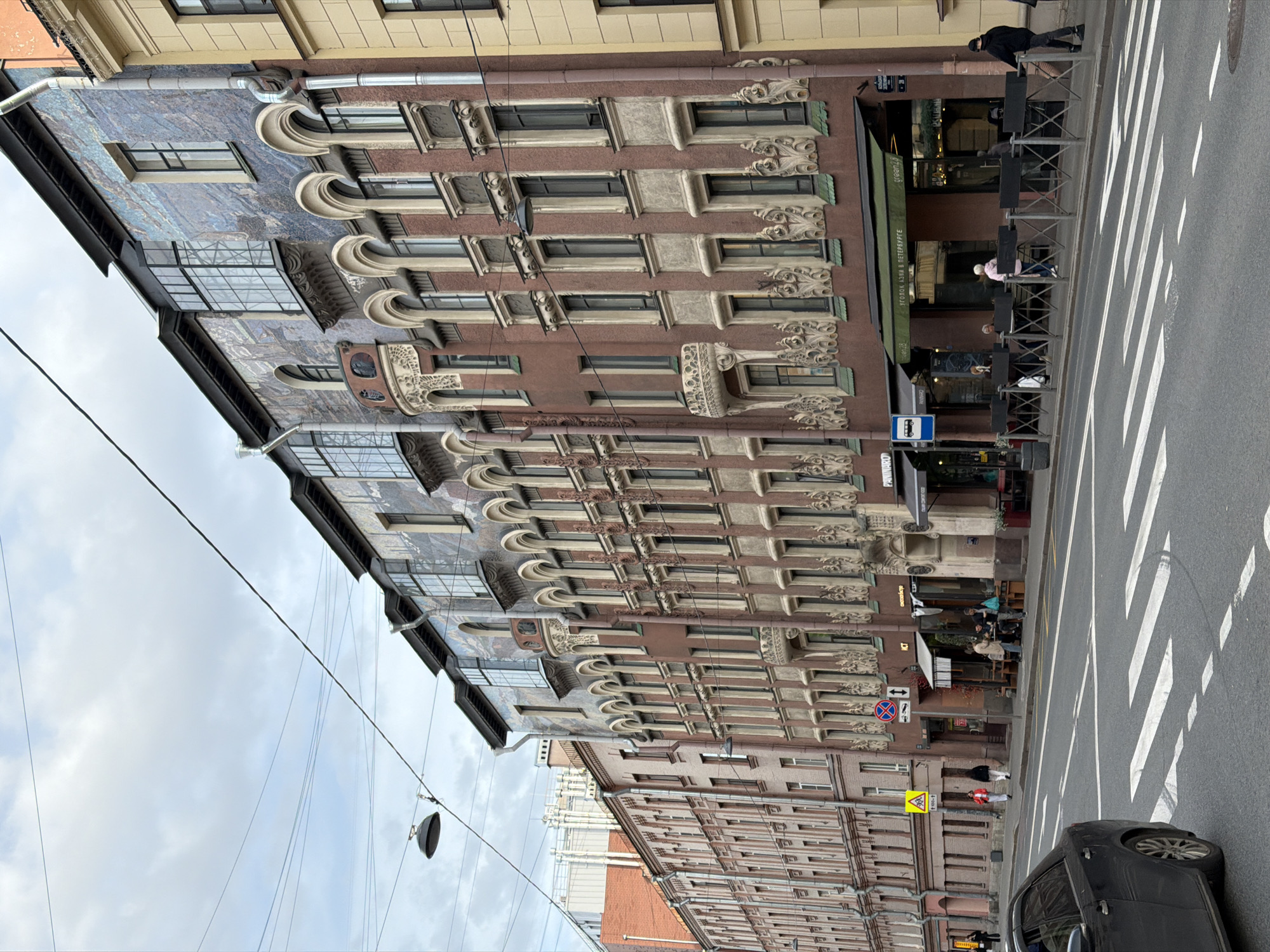 This image depicts a bustling street scene in Saint Petersburg, Russia, centered around an ornate, multi-story building. The building features a reddish-brown facade richly decorated with intricate window frames and decorative elements, possibly in an Art Nouveau style. The upper section of the building's facade displays a distinctive blue mosaic or tiled pattern. Drainpipes are visible running vertically along the building.
On the ground floor, several commercial establishments are present, marked by green awnings. One prominent awning displays the text "УГОЛОК АЗИИ В ПЕТЕРБУРГЕ" (Corner of Asia in St. Petersburg), and below it, possibly "Green Tea." Outdoor seating areas with black tables and chairs line the sidewalk, where several people are seen sitting, suggesting they are patrons of a cafe or restaurant. Other individuals are walking along the sidewalk, including one pushing a baby stroller.
The street in the foreground features a white zebra crosswalk. A black car is visible on the right side of the road. Overhead, numerous power lines crisscross against an overcast sky, indicating daytime conditions with moderate, diffused light.
Additional visible text and signs include a blue square bus stop sign with a white bus icon, a red circular "No Stopping" traffic sign, a yellow triangular warning sign with a black pedestrian symbol, and partially visible text for "Panna Cotta" on another storefront. The overall atmosphere suggests a typical day on a lively city street.