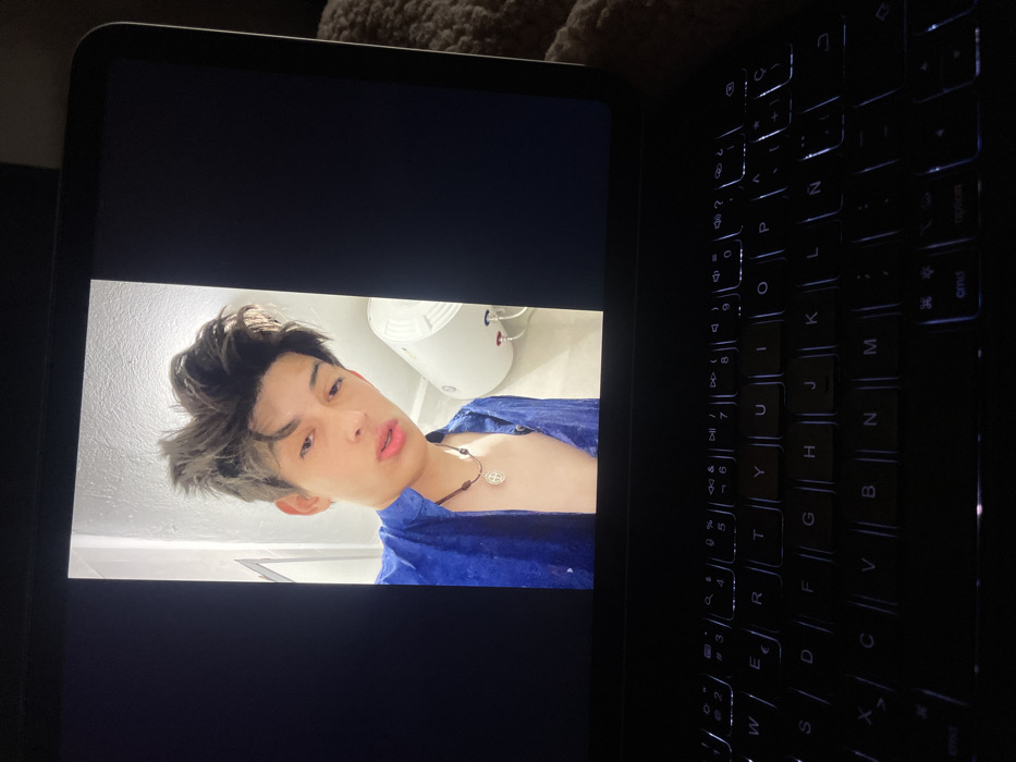 A laptop displays an image of a young man in a blue shirt with a necklace, facing the camera at an angle, with a white object in the background. The laptop's keyboard is partially visible on the right side, illuminated from underneath. The overall setting appears dimly lit, accentuating the laptop screen and keyboard.