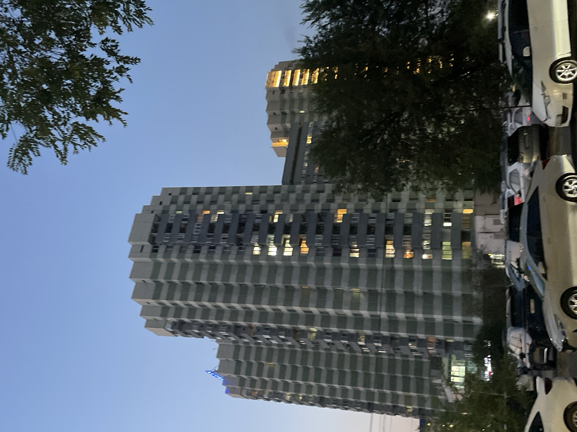 The image depicts an urban scene at dusk, likely in Almaty, Kazakhstan, given the architectural style of the apartment buildings. In the foreground, several cars are parked, suggesting a residential area. The buildings themselves are tall, multi-story apartment complexes with a distinctive pattern of light and dark grey horizontal bands. Many windows in the buildings are illuminated, indicating that residents are home. In the middle ground, more apartment buildings are visible, one of which has a more distinctly zigzagging roofline and brightly lit floors. The sky is a clear, twilight blue, transitioning from the evening light. A large tree with dark green foliage partially obscures the view of the buildings on the right side of the frame. The overall impression is a typical urban residential neighborhood settling in for the evening.