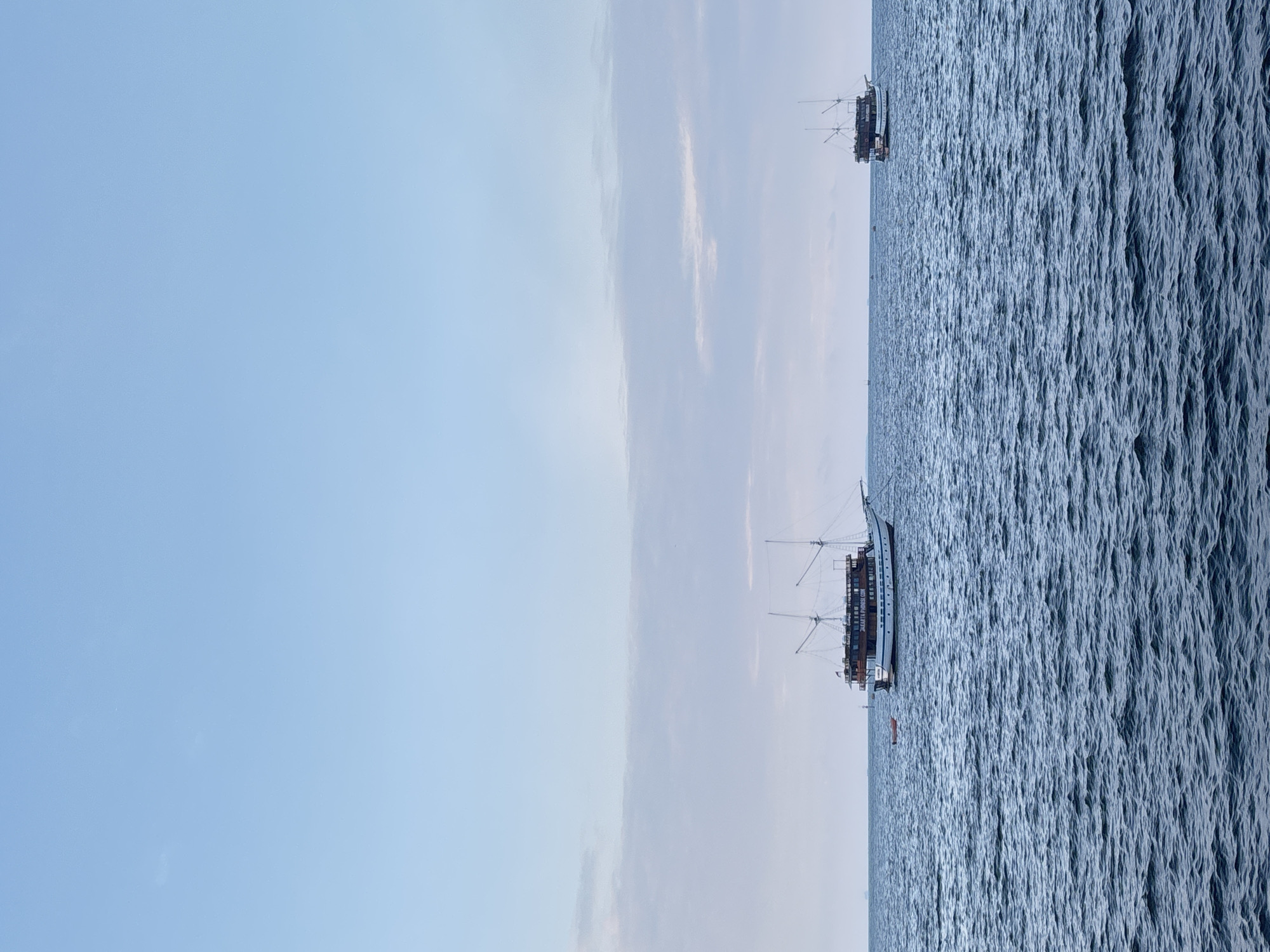 Two large boats, one more prominent in the foreground and the other in the background, sail on the open sea under a clear, pale blue sky with scattered clouds. The foreground boat is a traditional wooden sailing vessel with multiple masts and a dark superstructure, anchored near a small, orange dinghy. The background boat is similar in design but appears smaller and further away. The water is a deep blue with visible waves, suggesting a breezy day. The scene is tranquil and evokes a sense of maritime activity. This image likely depicts a scene off the coast of North Jakarta, Indonesia, a region known for its busy port and coastal waters.