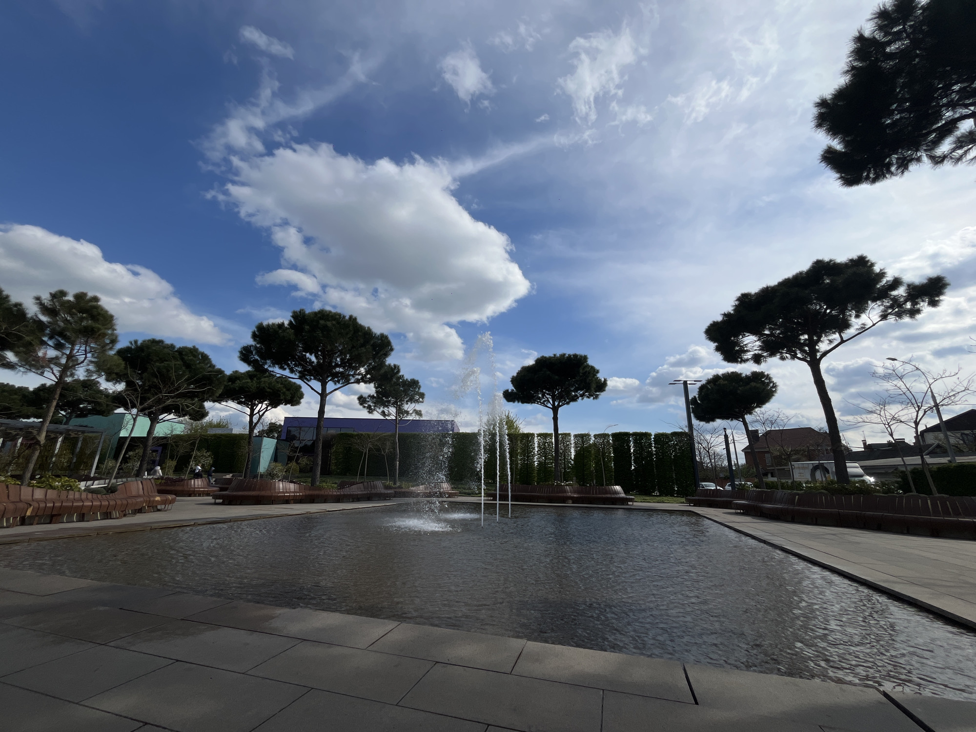 This image depicts a modern, well-maintained public park or plaza in Krasnodar, Russia, under a clear blue sky dotted with scattered white clouds. The central feature is a large, rectangular, shallow pool filled with dark water, from which several multi-stream fountain jets actively spray water upwards. The pool is bordered by light-colored, neatly paved pathways.
The park is furnished with numerous long, curved benches made of dark wooden slats, arranged in sections around the pool area, some forming circular seating arrangements. Several mature trees with dark green foliage and distinctive, often flat-topped or umbrella-like crowns are strategically placed, adding to the aesthetic. Behind these trees, a tall, dense, and meticulously trimmed dark green hedge provides a structured backdrop.
In the mid-ground, parts of modern buildings with angled architectural elements are visible. Further in the background on the right, a street scene unfolds with traditional-style houses and a white delivery truck, indicating an urban setting. A few indistinct figures of people can be seen seated on benches or walking in the distance, suggesting leisure activity. The overall atmosphere is peaceful and inviting, captured during what appears to be a bright, sunny day. No discernible text is visible within the image.
