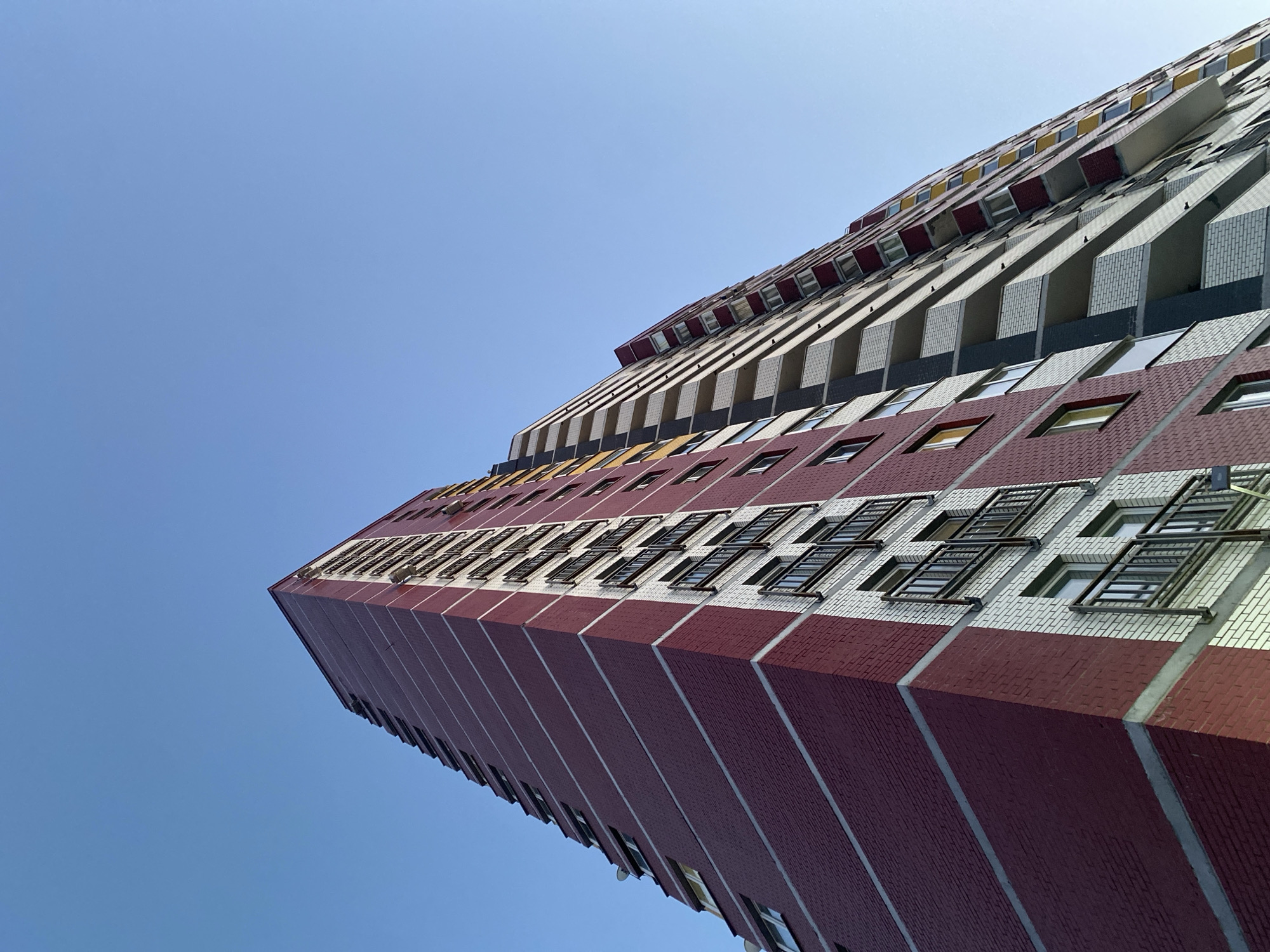 The image shows a low-angle shot of a tall, multi-story building in Izhevsk, Russia. The building has a distinctive facade with alternating horizontal bands of red brick and lighter-colored brick or paneling. Windows are arranged in rows, some with window guards. The building extends upwards at an angle into a clear blue sky. The architecture is modern and utilitarian. The building appears to be residential.