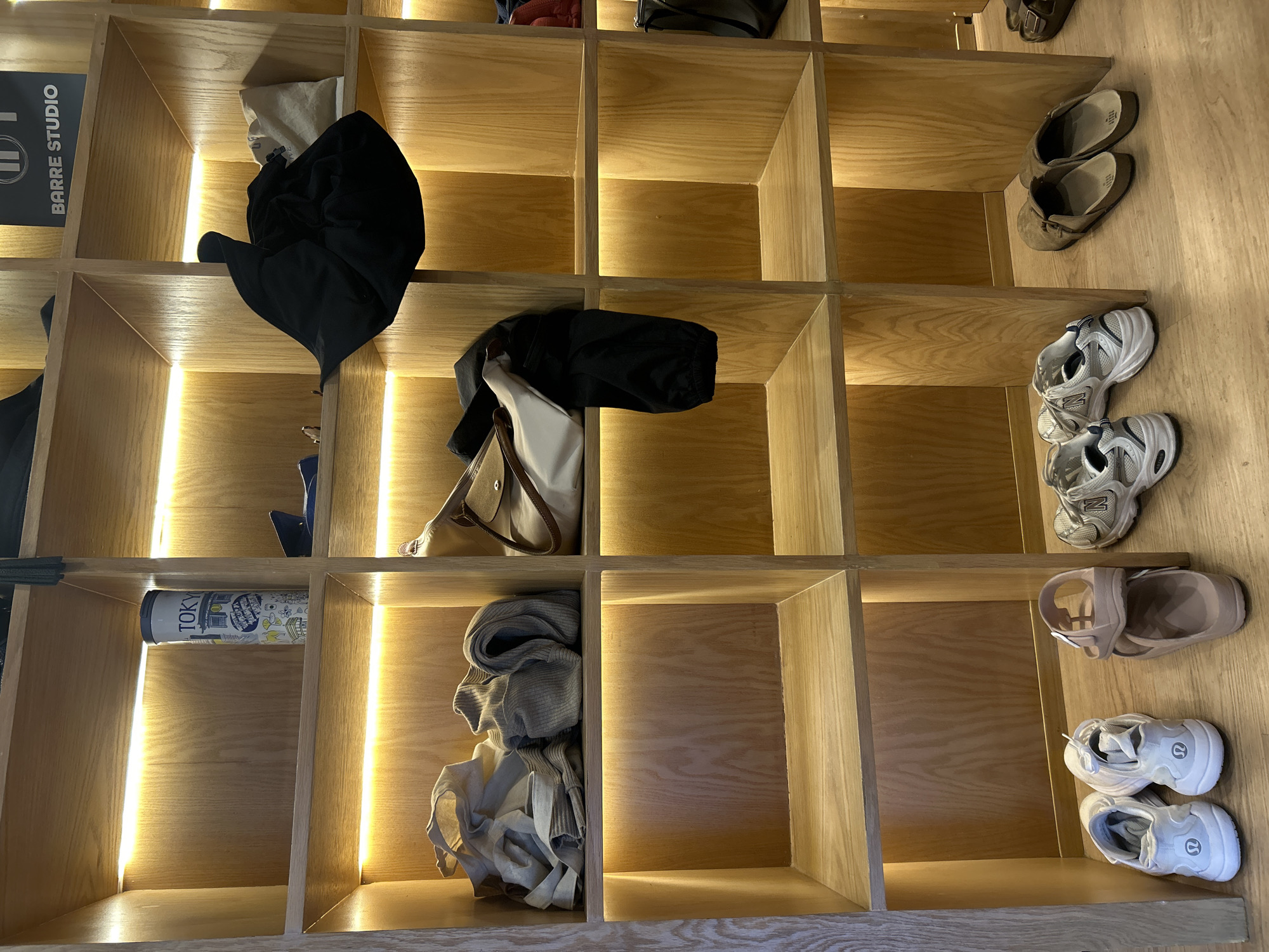 The image captures an interior scene, likely a changing room or locker area within a fitness studio, specifically a "BARRE STUDIO" as indicated by a black sign with white text in one of the cubbies. The setting features a large, light wooden shelving unit, creating a grid of individual square compartments or cubbies. Each cubby is illuminated along its vertical sides by warm LED strip lighting, casting a soft glow and highlighting the wood grain. The floor beneath the shelves is also made of wood.
Various personal items are stored within or next to these cubbies. In the top left, a black garment and a light bag share a cubby. Another cubby further along the second row contains a black jacket or hoodie draped over a light-colored handbag with brown straps. On the floor to the right, a pair of brown slip-on shoes or sandals, a pair of grey and white New Balance athletic shoes, a pair of light pink open-toe sandals, and two pairs of white Lululemon athletic shoes are neatly placed. One cubby contains a white cylindrical container with blue and black graphics, labeled "TOKYO." Another cubby in the bottom row holds folded light-colored clothing. Many cubbies remain empty, suggesting active use of the facility. The scene is well-lit and appears clean and organized.