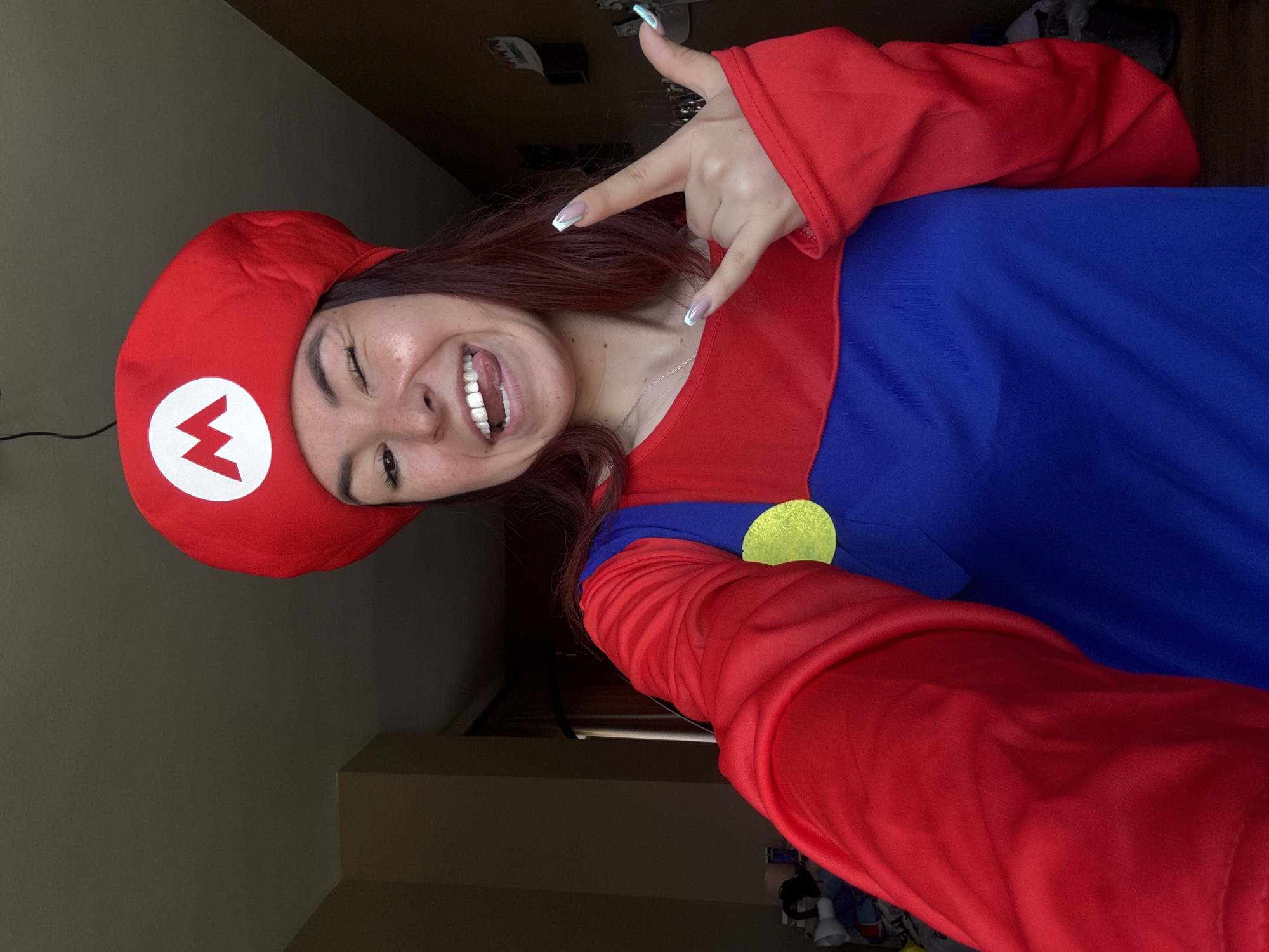 A person dressed in a Mario costume is taking a selfie indoors. The person is wearing a red hat with a white circle containing a red "M," a blue shirt with a large yellow button, and red sleeves. The person is making a rock-and-roll hand gesture with their left hand, winking, and smiling with their tongue slightly out. The background is a neutral color wall and a dark-colored wooden surface with other objects in the background, not clearly identifiable. The image does not contain details that suggest a location in Ruse, Bulgaria.