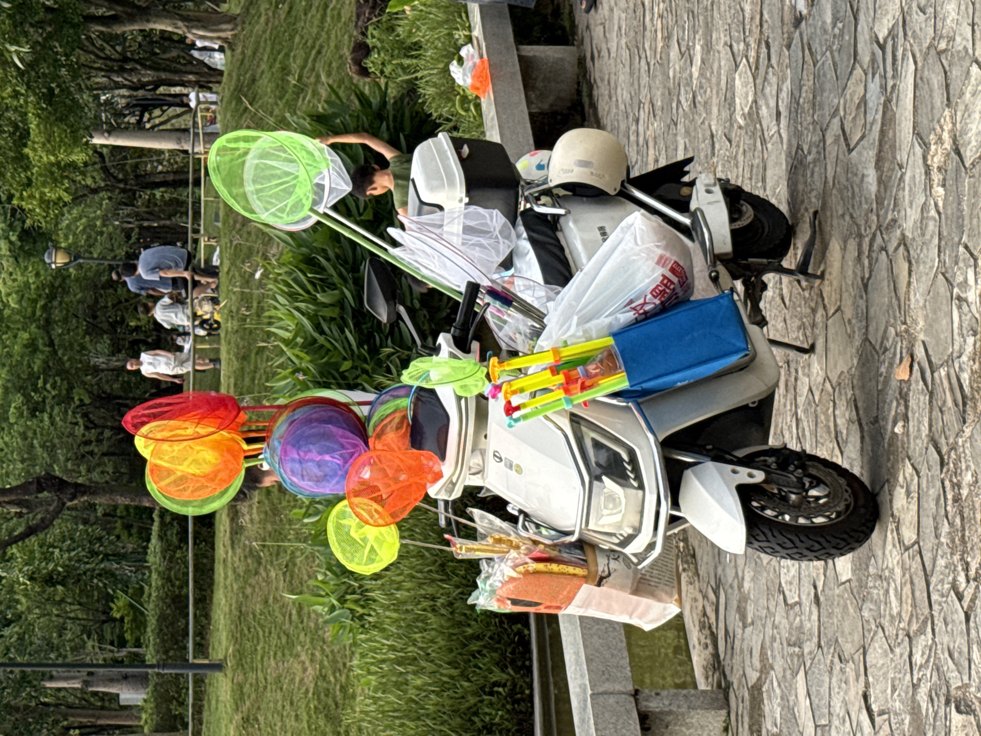 A white electric scooter is parked on a cobblestone path in a park, likely in Huizhou, China, as suggested by contextual clues. The scooter is heavily laden with various items, primarily colorful butterfly nets of different sizes and hues, including red, orange, yellow, green, blue, and purple. Several long, yellow, and red plastic poles, possibly for the nets, are also attached to the scooter. A blue bag hangs on the left side, containing a few orange items. A white helmet is secured to the back of the scooter. In the background, a lush green park area with trees and a grassy slope is visible. A few people are present in the background, including a child in a green shirt holding a large green net, and other individuals sitting and walking near a railing. The overall scene suggests a vendor selling park-related items, such as nets for catching insects, to park visitors.
