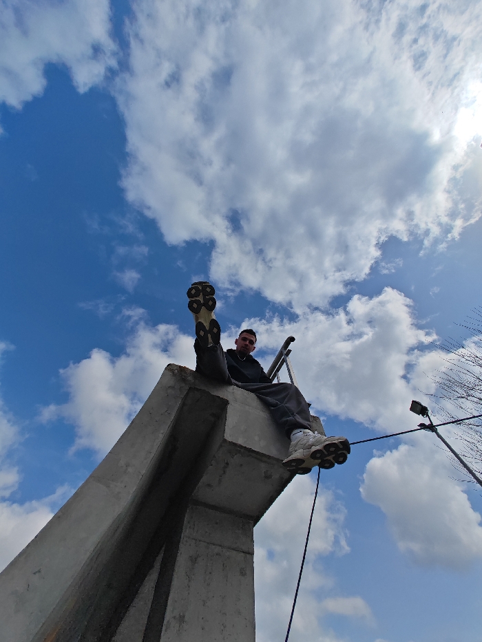 A young male is depicted seated on a large, angular concrete structure, identified as part of the Monument to 1300 Years of Bulgaria in Shumen, Bulgaria. He wears a dark hoodie, dark sweatpants, sunglasses, and light-colored chunky sneakers. The male is positioned with his legs extended forward, his left leg raised higher, prominently displaying the sole of his shoe, and he looks directly into the camera. A dark metal railing is visible behind and to his right. A dark-colored cable or rope extends vertically downwards from the concrete structure near his right side. The background consists of a bright blue sky with scattered white cumulus clouds. The sun is partially visible in the upper right, creating a bright glare. To the right, a bare tree and a utility pole with a streetlight fixture are visible.