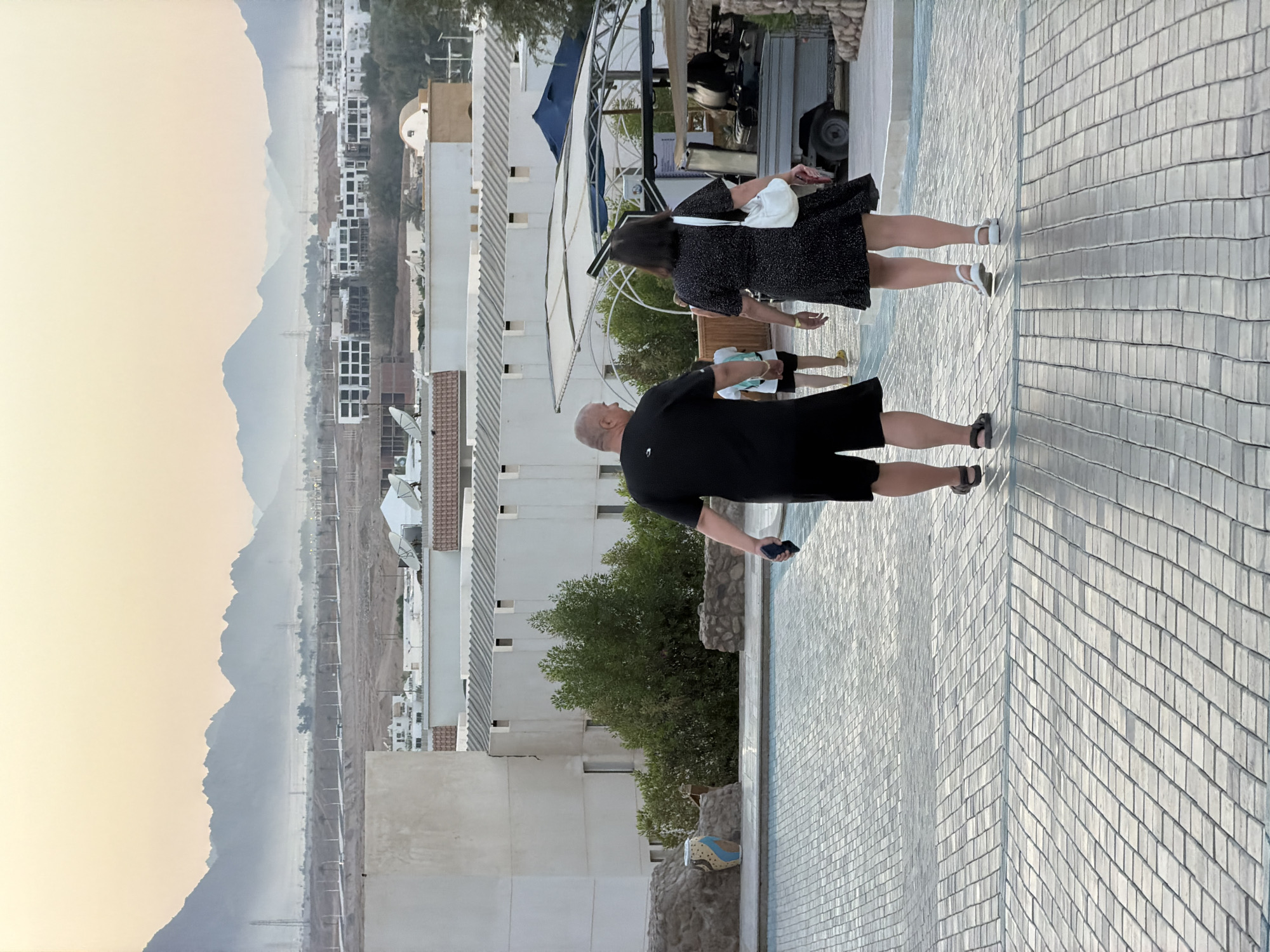 In Sharm El Sheikh, Egypt, an outdoor scene unfolds on a tiled walkway. Two adults, a man and a woman, are walking away from the camera. The man, on the left, is wearing a black t-shirt and black shorts, holding a dark object in his right hand. The woman, on the right, is wearing a black dress with a white purse slung over her shoulder. Behind them, other figures are visible, including a child carrying a backpack. The walkway curves upwards and leads towards a resort or hotel complex with white buildings and some greenery. In the background, a hazy mountain range is silhouetted against a pale, overcast sky, suggesting an early morning or late afternoon timeframe. The general environment appears to be a well-maintained tourist area.