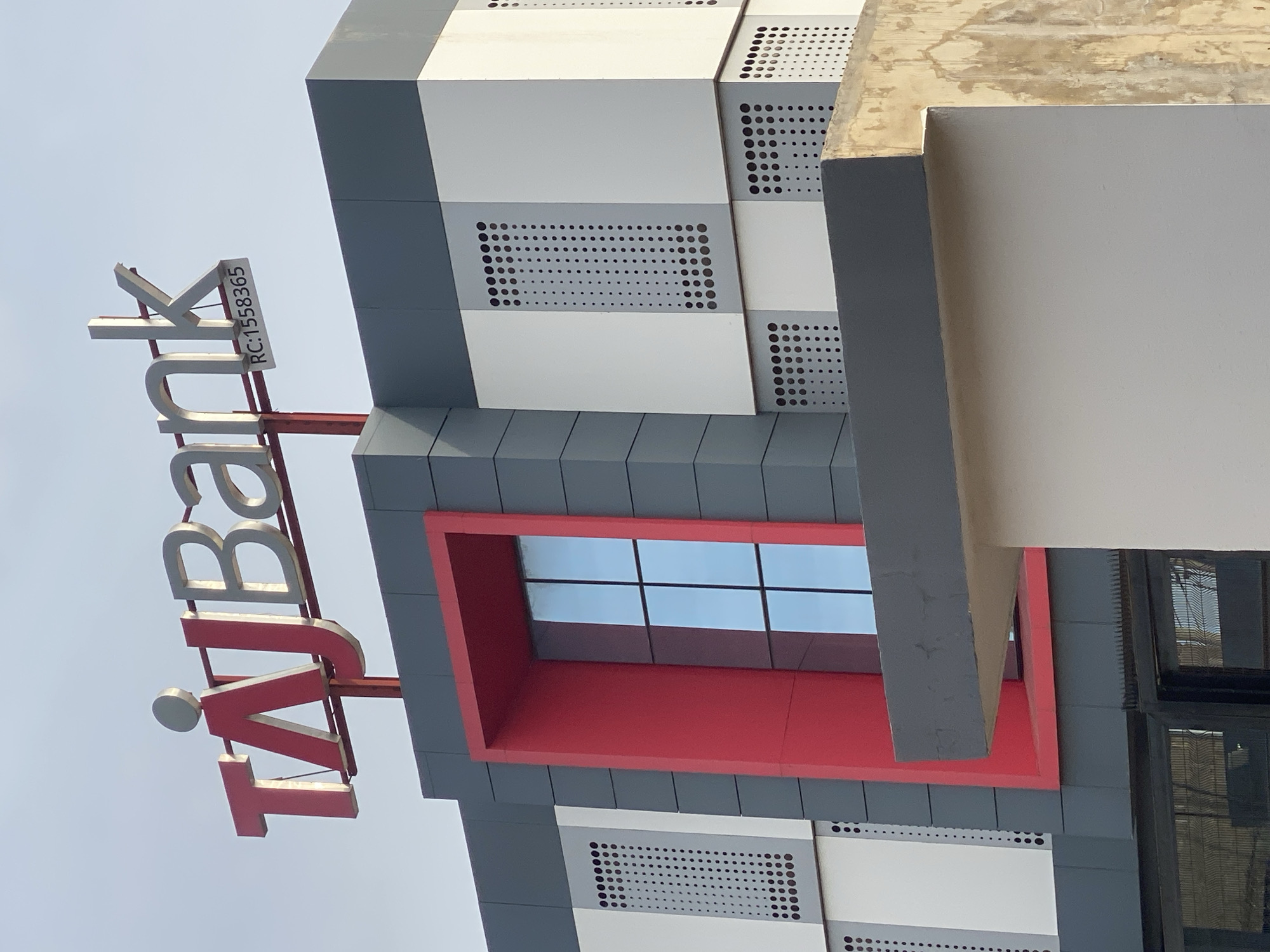 The image shows the exterior of the TAJ Bank building in Sabon Tasha, Nigeria. The building features a modern architectural design with a prominent red and grey color scheme. The TAJ Bank logo, in red and silver, is prominently displayed at the top, along with the registration number "RC:1558365". The building has a large, multi-paned window framed in red. The facade is composed of alternating grey and white panels, some of which have perforated patterns. The foreground shows a section of a light-colored wall and a dark metal gate. The sky is visible in the background.