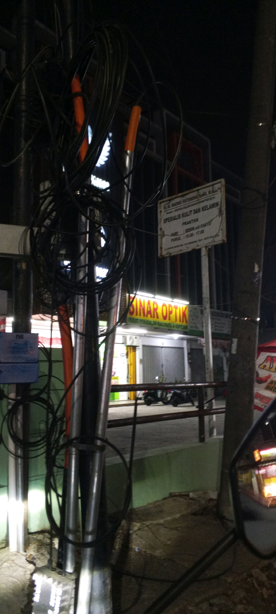 The image depicts a nighttime street scene in Cibinong, Indonesia, with a focus on a tangled mess of electrical wires and cables attached to a pole in the foreground. Behind the wires, a brightly lit sign for "SINAR OPTIK" is visible, indicating a business that sells eyeglasses and contact lenses. Further back, the facade of the optical shop can be seen, with its entrance and windows illuminated. Several motorcycles are parked in front of the shop, suggesting it is a popular establishment. The environment appears to be an urban area, with the presence of streetlights and buildings. The overall impression is one of a typical Indonesian street scene at night, characterized by a mix of commercial activity and urban infrastructure.