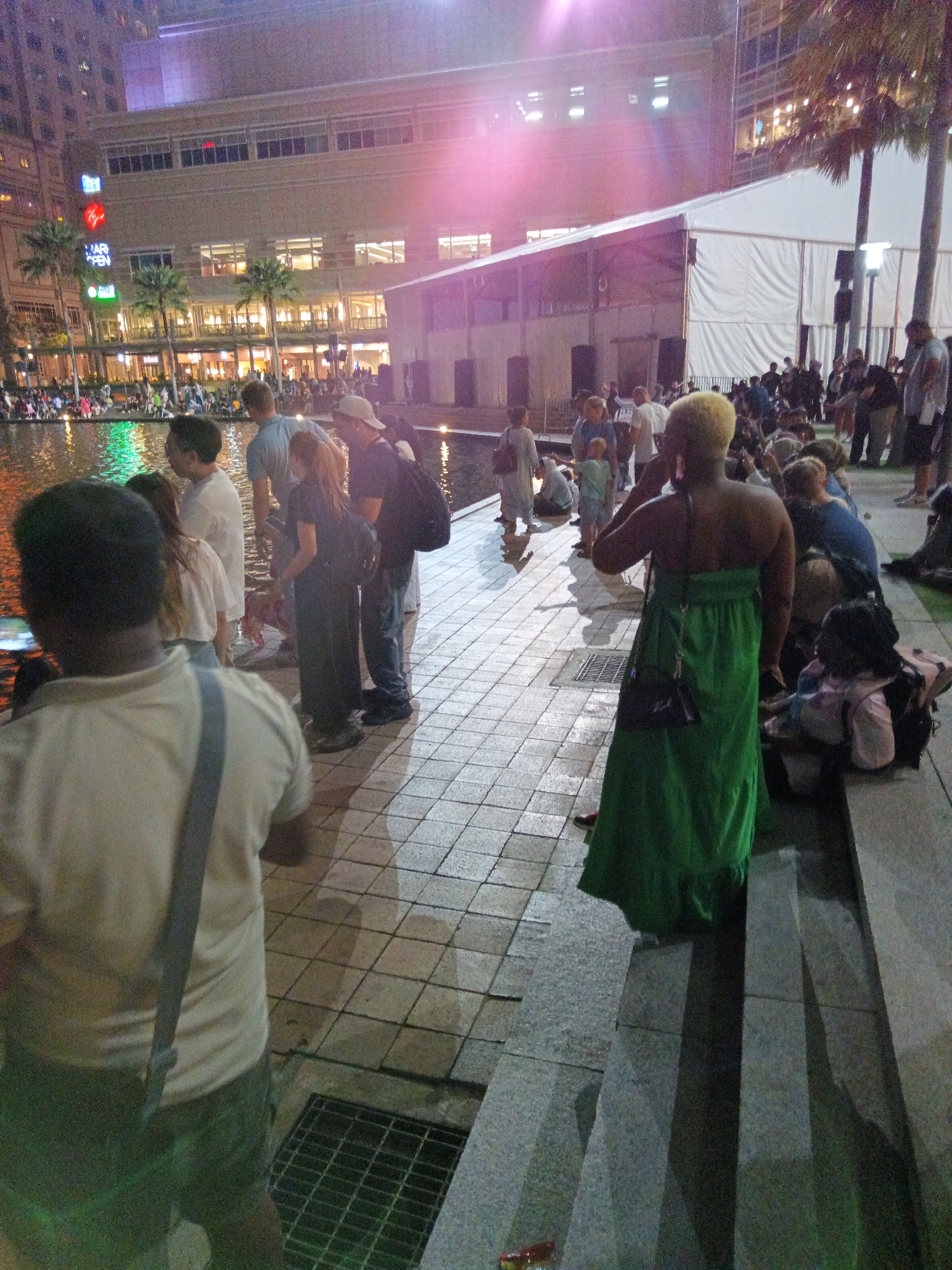 The image depicts an outdoor evening scene in Kuala Lumpur, Malaysia, likely near a waterfront area in front of a large building with illuminated windows. Several palm trees are visible, and there is a large white tent structure in the background. A group of people are gathered, with some standing by a body of water that reflects green light. In the foreground, a person in a beige shirt and green shorts is facing away from the camera, holding a phone. To the right, a person in a long green dress is standing and facing away from the camera, with a black bag slung over their shoulder. Other individuals are seen in various states of interaction, including talking and observing. The ground is paved with tiles, and there are concrete steps. The lighting suggests it is dusk or nighttime, with artificial lights contributing to the ambiance.