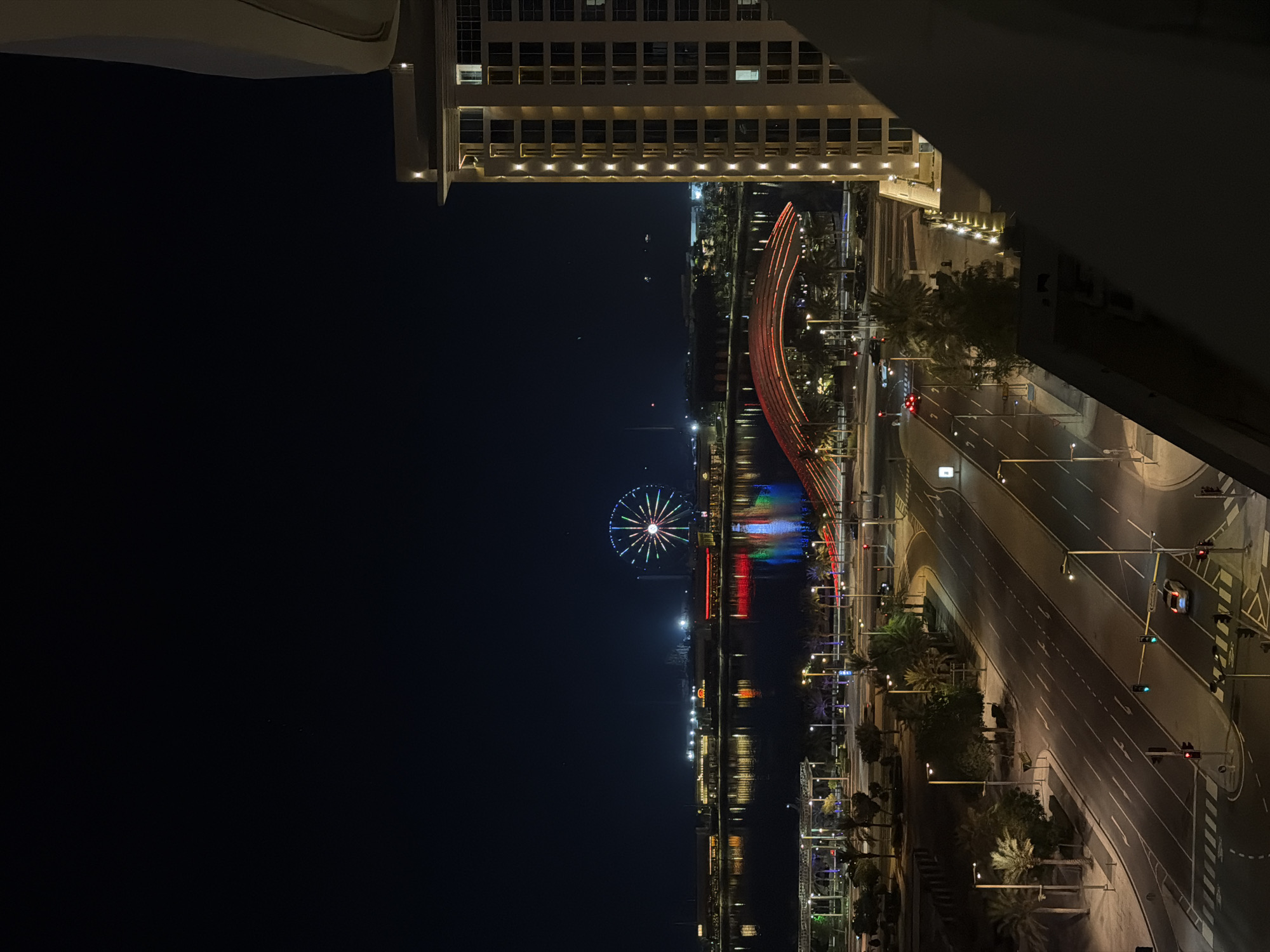 An aerial nighttime shot captures a city scene with a large Ferris wheel illuminated with colorful lights in the distance. A body of water reflects the lights of buildings and the Ferris wheel. In the foreground, a multi-lane road with streetlights and palm trees borders a modern building on the right. Cars are visible on the road and at an intersection with traffic signals. The scene conveys a vibrant urban atmosphere.