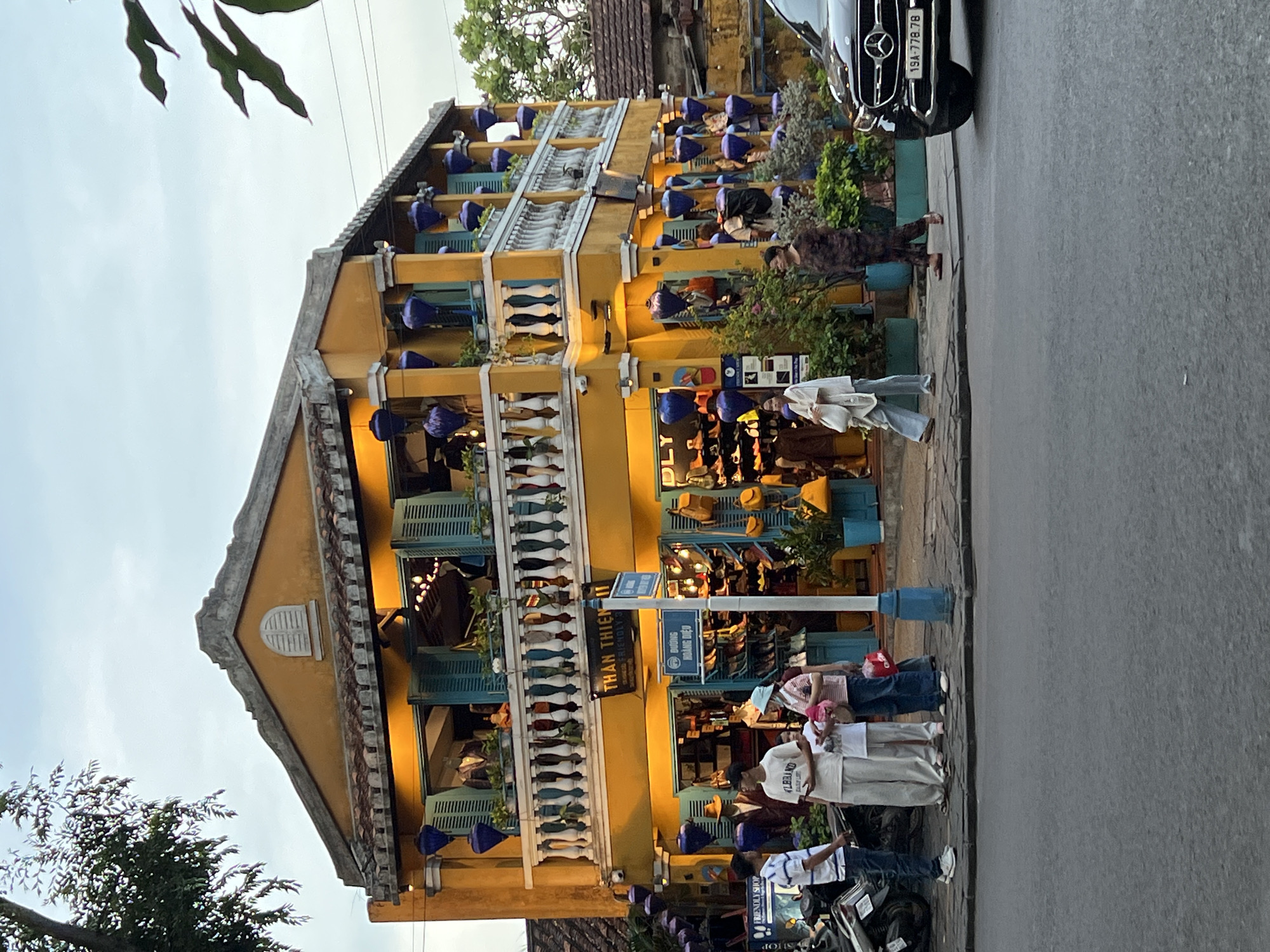 This is a medium shot, eye-level, outdoor photograph of a yellow building with blue shutters and a balcony on a street in Hội An Ward, Vietnam. The building appears to be a shop. It is decorated with numerous blue lanterns. The shop's name, "THAN THIEN FRIENDLY," is visible on a sign.
In the foreground, a black Mercedes-Benz car is parked on the right side of the street. The street is paved, and a sidewalk runs along the front of the building.
Several people are visible on the sidewalk. On the left, three individuals stand near a motorcycle. Two men are wearing casual clothing, and one is wearing a white shirt with blue stripes, jeans, and white sneakers. A young person in a white t-shirt and beige pants is standing next to them. A woman in a pink and white striped shirt and blue jeans, holding a red bag, is also present.
Further to the right, a woman in a white outfit walks past the shop. Another woman, wearing a dark patterned outfit, is walking towards the right of the frame.
The building has two stories. The ground floor is filled with merchandise, including shoes and bags displayed in the windows and on shelves. The second story features a balcony with a white railing, also displaying items for sale. The building's facade is adorned with potted plants and lights. The sky is overcast.