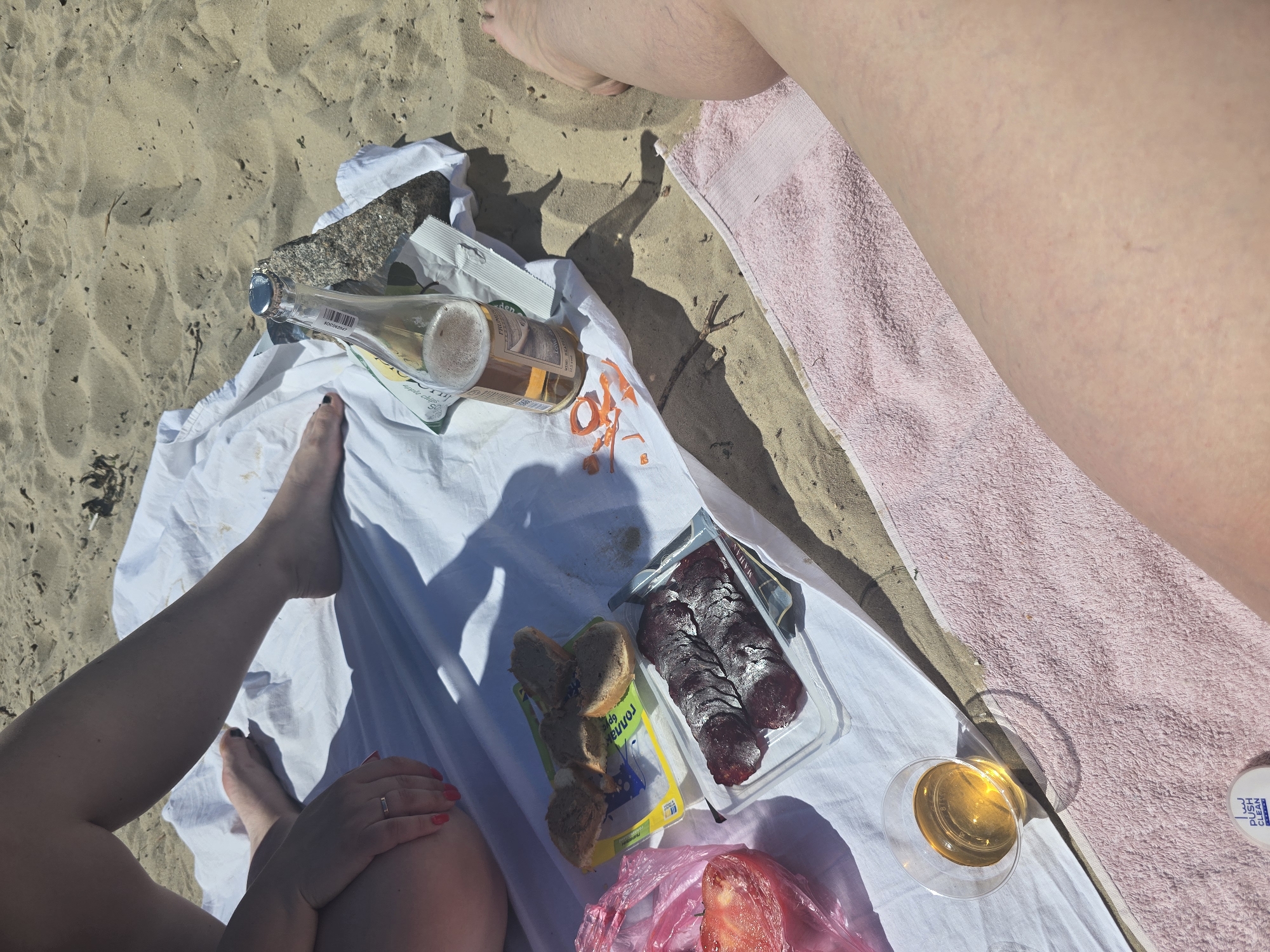 The image shows a picnic on a sandy beach, likely in Kyiv, Ukraine, given the context. The scene is viewed from above, focusing on a white blanket spread on the sand. Two individuals are present, partially visible, with legs and feet shown. One person's legs are on the right side of the frame, resting on a pink towel. The other person's legs and feet are on the left side of the frame, positioned on the white blanket.
On the blanket, there is a picnic spread. A bottle of sparkling wine or beer is open and appears to be partially consumed, with foam visible. Next to it, there is a package of sliced cured meat, possibly sausage. There are also some pieces of bread, a pink plastic bag containing what looks like a sliced tomato, and a glass of amber-colored liquid, likely the same beverage from the bottle. A small white container with blue text is also visible, and a piece of rock is near the bottle.
The time of day appears to be sunny and bright, indicated by the strong shadows cast by the objects and people. The weather seems warm and pleasant, suitable for a beach picnic. The overall mood suggests a relaxed and enjoyable outdoor activity, perhaps a summer day. The image captures a casual moment of enjoying food and drink by the water.