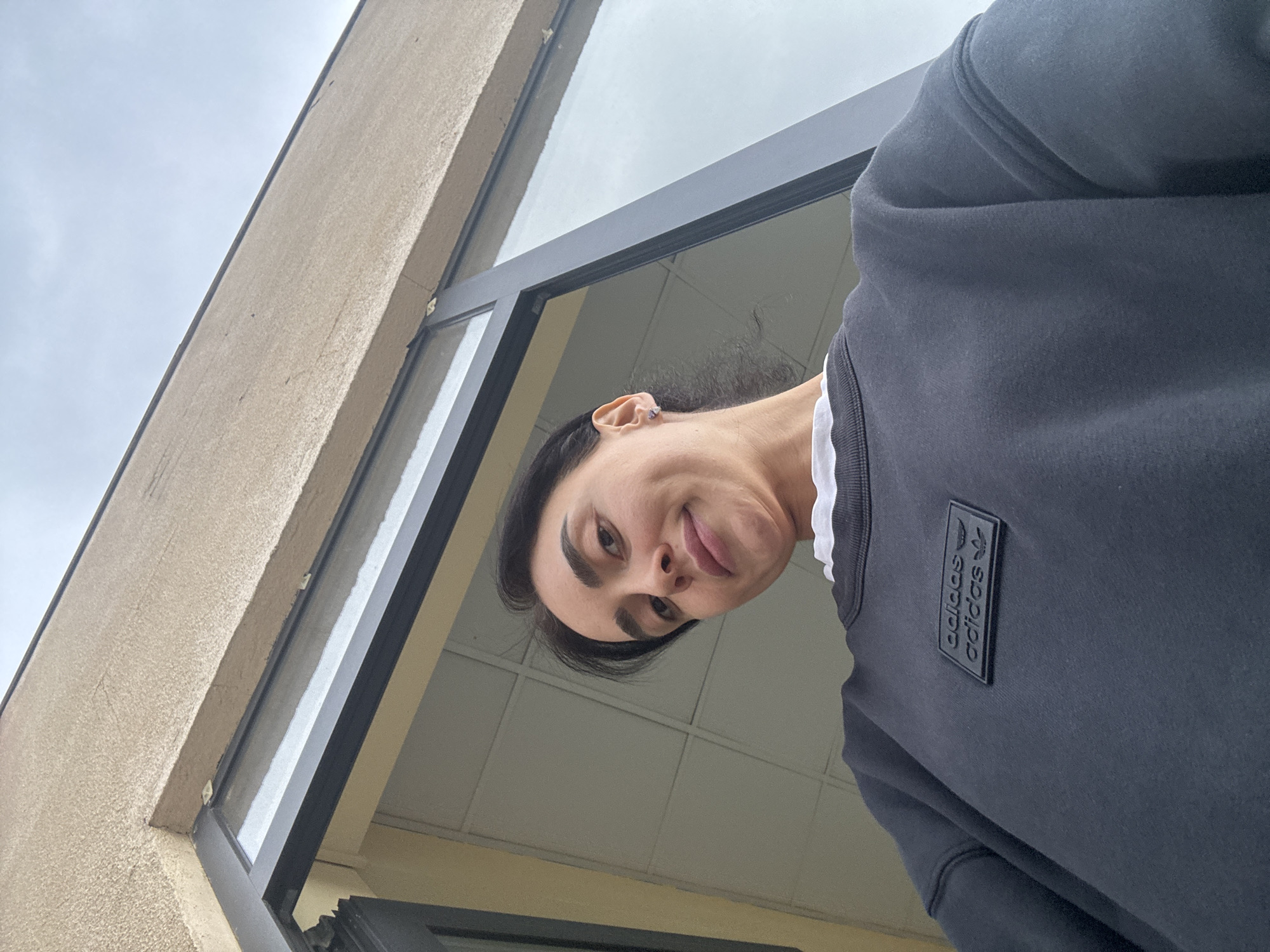 A woman in a black Adidas sweatshirt looks down at the camera from a window. The window frame is black and the building exterior is beige. The location appears to be Tbilisi, Georgia, based on architectural styles and urban environment.