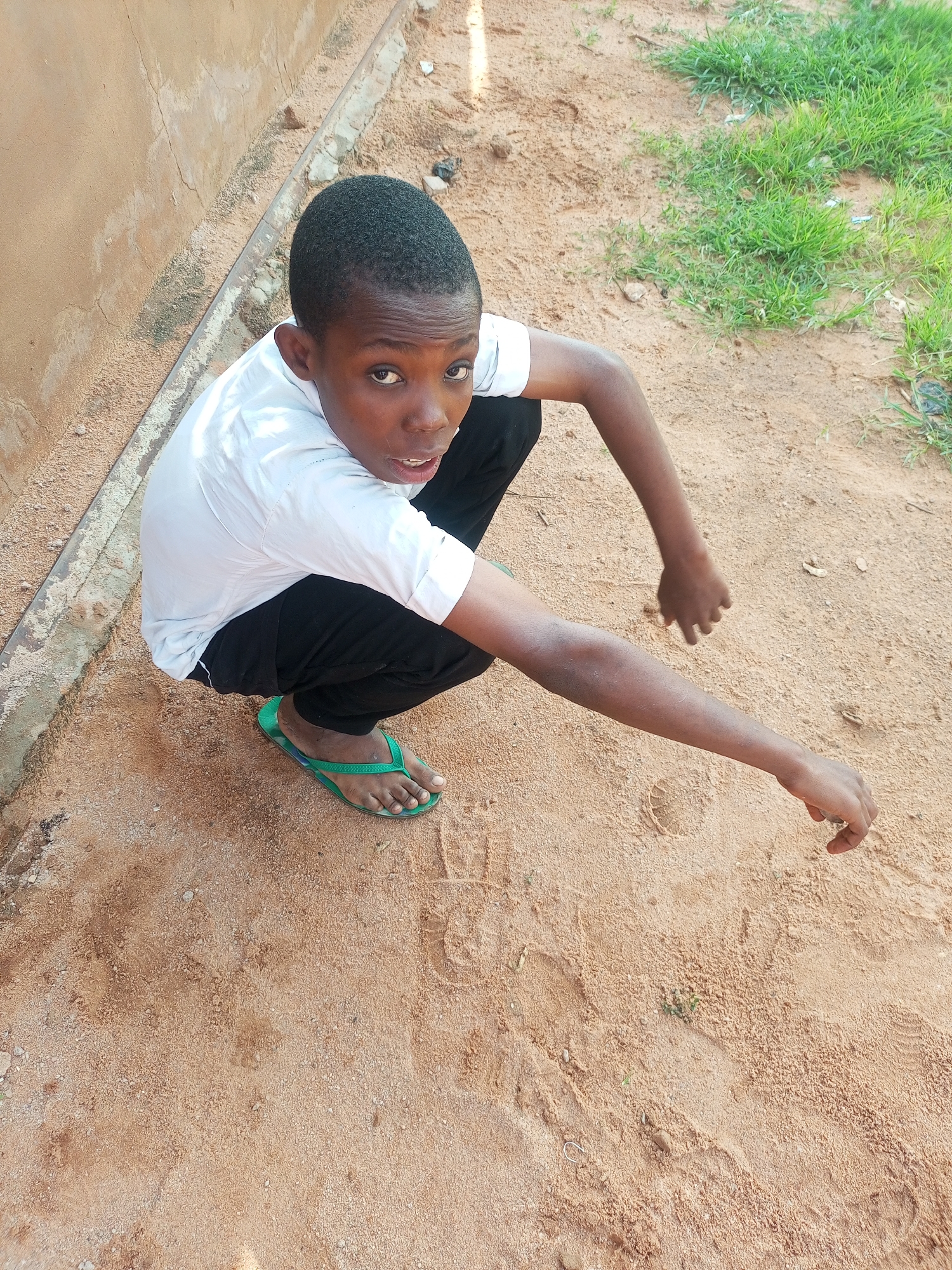 A young Black male is crouching on sandy ground next to a wall. He is wearing a white t-shirt and black pants, with green flip-flops on his feet. His hands are resting on the ground in front of him. In the background, there is some grass. The lighting suggests it is daytime.
