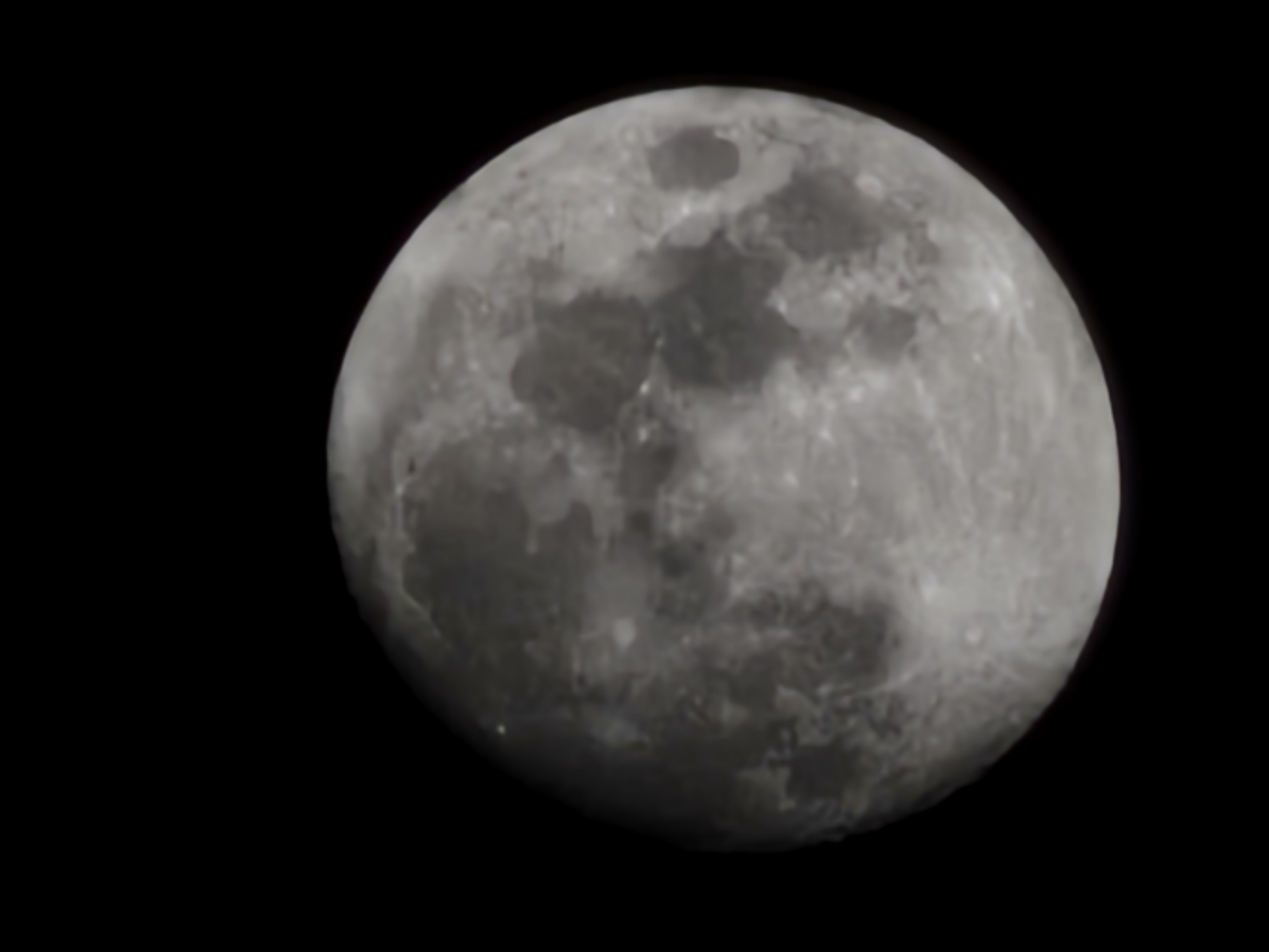 The image depicts the moon against a black background. It shows a full or nearly full moon with craters and variations in surface texture visible. The moon is gray in color. The image does not contain any information about the location in Esch-sur-Alzette, Luxembourg, people, actions, interactions, or environment.