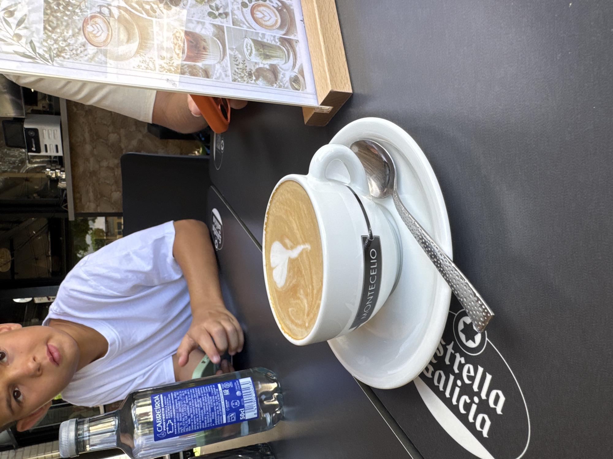 A young boy in a white t-shirt sits at a dark table outdoors, his gaze directed slightly upward and to the left. A partially filled bottle of Cabreiroá water is on the table to his left, and his hands are resting on his lap. In the foreground, a white ceramic cup with the name "MONTECELIO" printed in black sits on a saucer. The cup contains a latte with foam art. A silver spoon rests on the saucer. To the right of the cup is a wooden stand displaying a menu with several images of drinks, likely at a cafe in La Vila Joiosa, Spain.