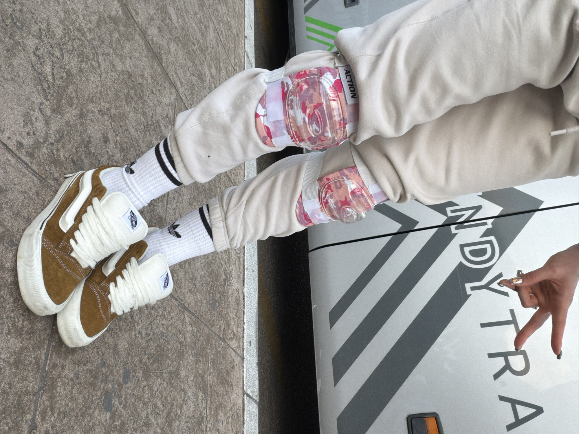 A full shot captures a person wearing khaki-colored sweatpants, knee pads, white socks with black stripes, and brown suede Vans sneakers. The person stands on a weathered, grey tiled sidewalk, partially obscured by a white vehicle with grey stripes and the word "NDYTRA" printed on it. The vehicle is positioned beside the person, the person's right arm resting on the vehicle. The location is Budva, Montenegro.