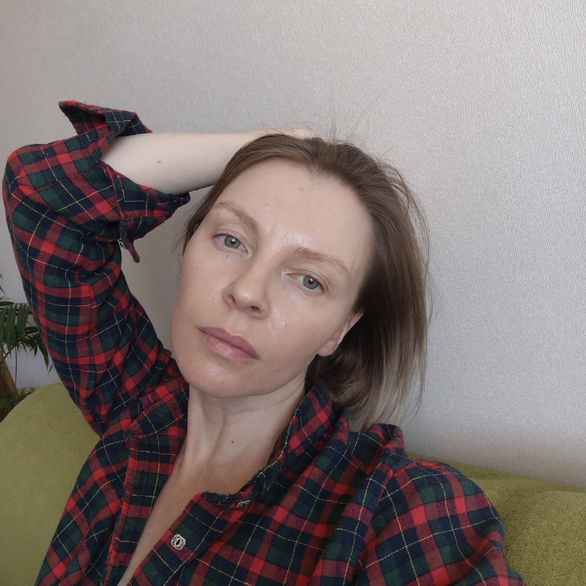 A woman with light skin and short, light brown hair is posing for a selfie. She has light green eyes and is wearing a red, green, and blue plaid flannel shirt. Her left arm is raised and resting on her head. She is sitting on a green couch in front of a beige wall. A potted plant is visible in the background. The location in the city of Chaikovsky, Russia is not discernible from the image.