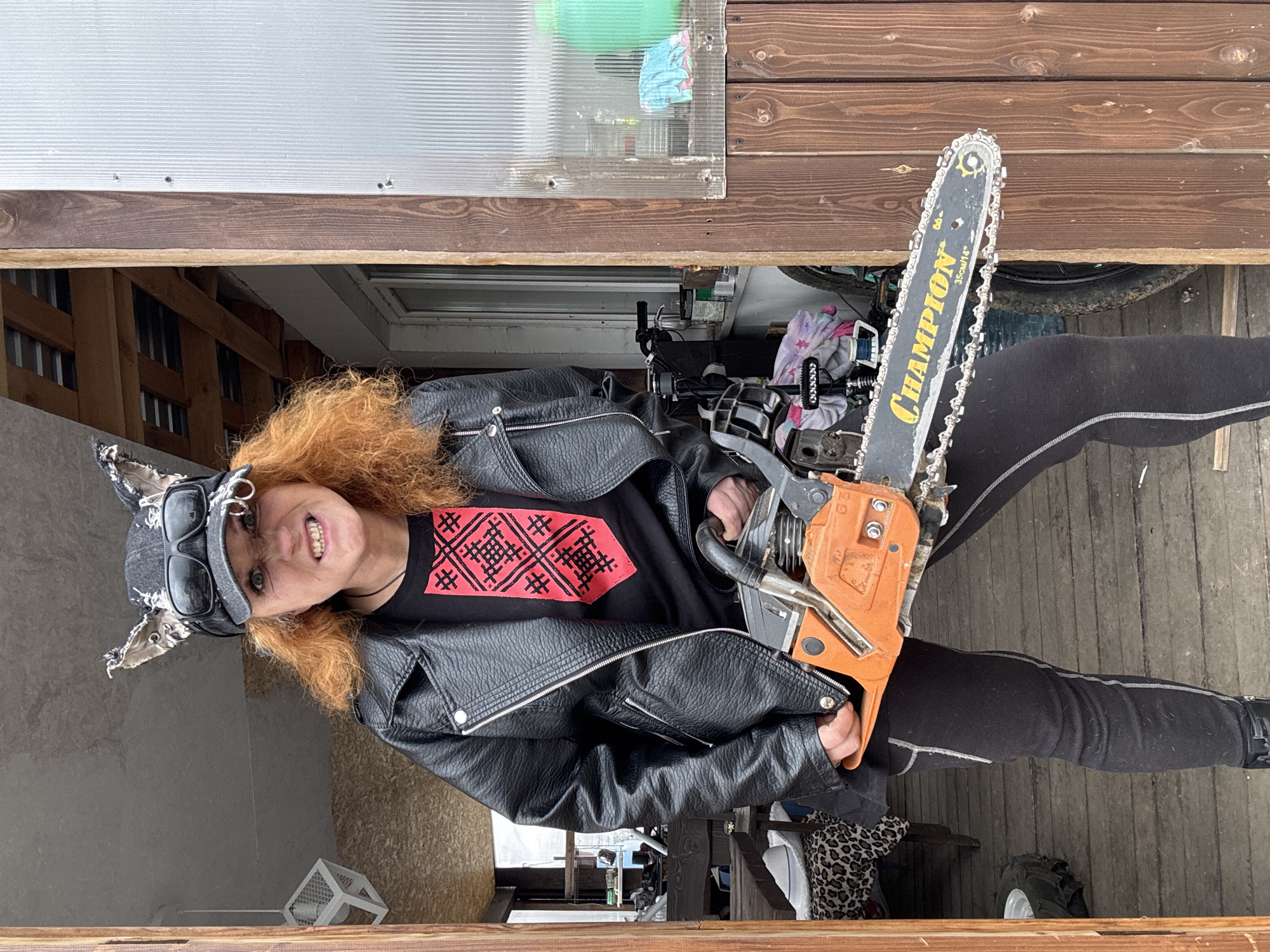 The image, originally rotated 90 degrees clockwise, depicts a person with bright, curly red-orange hair, standing outdoors in Chaikovsky, Russia. The subject, a woman, is shown from the waist up, smiling broadly with her teeth visible and looking directly at the camera. She wears a black leather jacket over a black t-shirt featuring a prominent red geometric pattern. On her head, she has a dark cap with cat-ear-like adornments, with a pair of dark sunglasses resting on top.
She is holding an orange and black chainsaw, its blade pointing upwards. The brand name "CHAMPION" is clearly visible in yellow lettering on the black blade, along with the measurement "35cm/14". The background consists of a mix of wooden panels and light-colored corrugated material, suggesting a shed, garage, or outdoor structure. Various other objects are dimly seen behind her, including what appears to be a bicycle wheel and some patterned fabric. The lighting is bright and even, indicating daytime. The subject's expression is one of confidence and a hint of playful intensity.