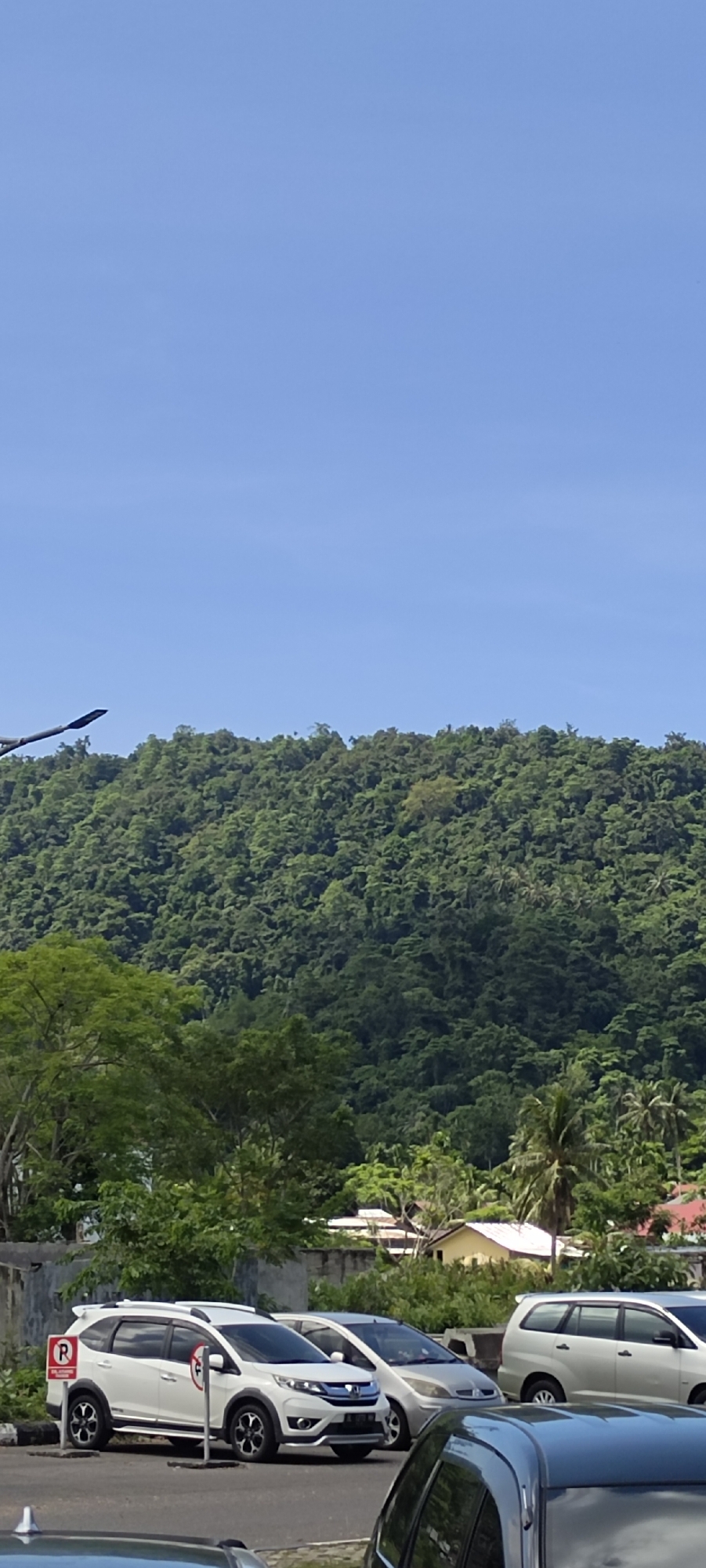 The image shows a view from a parking lot looking towards a lush, tree-covered mountain. Several parked cars are visible in the foreground, including a white SUV with a red "P" parking sign next to it. In the background, houses with orange and yellow roofs are scattered among the trees at the base of the mountain. The sky is a clear, bright blue. The scene appears to be in a tropical or subtropical location, consistent with the city of Sabang, Indonesia, which is known for its natural beauty and mountainous terrain. The lighting suggests it is daytime, likely late morning or early afternoon due to the bright sun and clear sky.