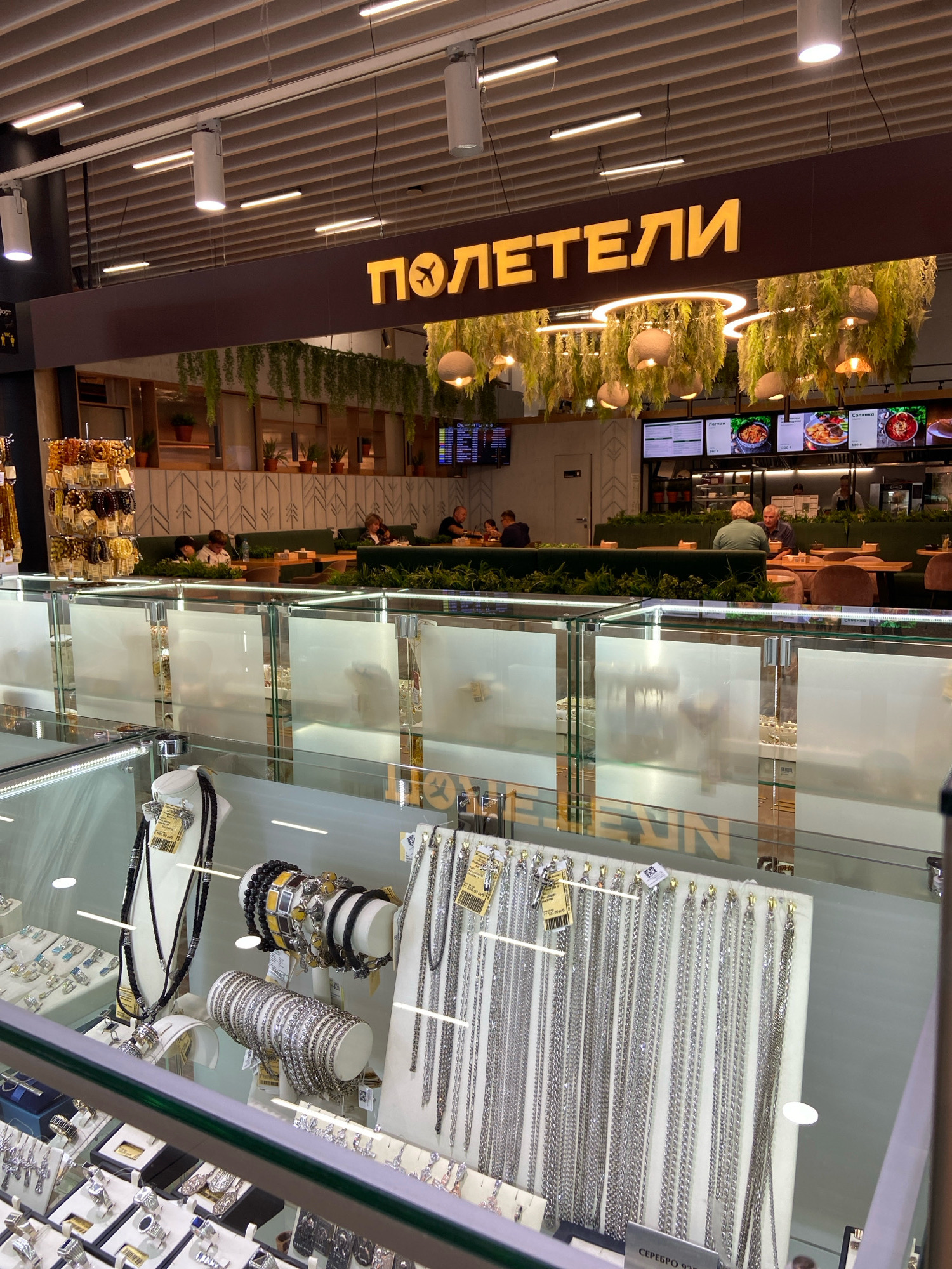 The image shows the interior of a jewelry store and a food establishment. The foreground features glass display cases filled with various jewelry items, including necklaces, bracelets, and rings. In the background, a food establishment with tables and seating is visible, where several people are seated and interacting. A prominent sign above the food establishment reads "ПОЛЕТЕЛИ" with an airplane logo, indicating a brand or name. The overall environment suggests an indoor commercial space, possibly a shopping mall or an airport terminal, given the presence of both a jewelry store and a food service area. The style of the interior suggests a modern design with a focus on lighting and decorative elements like hanging plants. It is not possible to determine the specific location within the city of Ob, Russia based solely on this image.