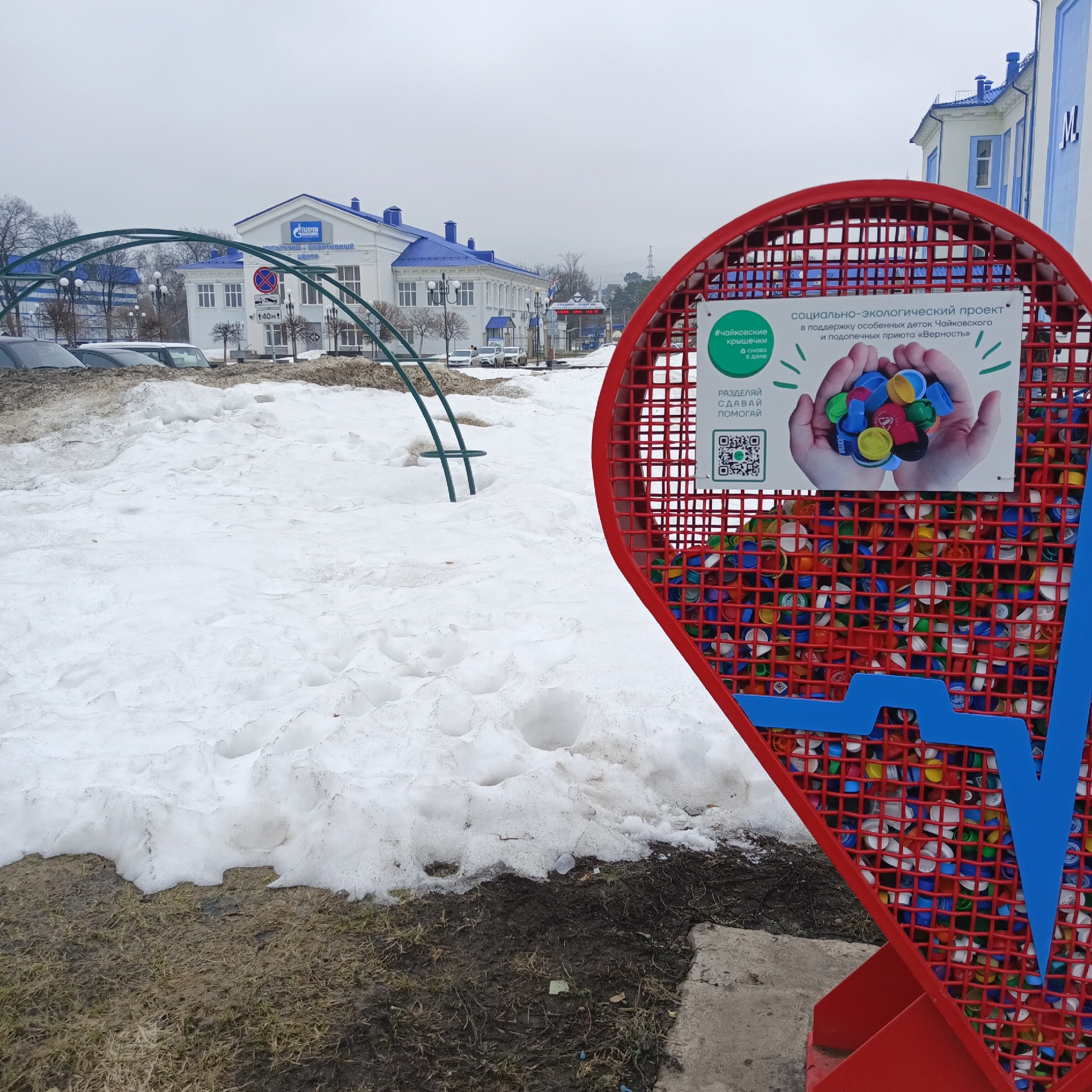 The image shows a street scene in the city of Chaikovsky, Russia. The foreground features a red metal structure, resembling a tennis racket, filled with colored plastic bottle caps. A sign on the structure promotes a "social-ecological project" to support children from Chaikovsky and a local shelter. The sign includes the text "#чайковские крышечки" (Chaikovsky lids).
Behind the structure is a snow-covered area with patches of bare ground. In the background, there are buildings, including a white one with blue accents and a sign reading "Газпром Культурно-спортивный центр" (Gazprom Cultural and Sports Center). Other visible objects include a metal archway, cars parked along the street, and a traffic sign indicating a weight limit. The sky is overcast.